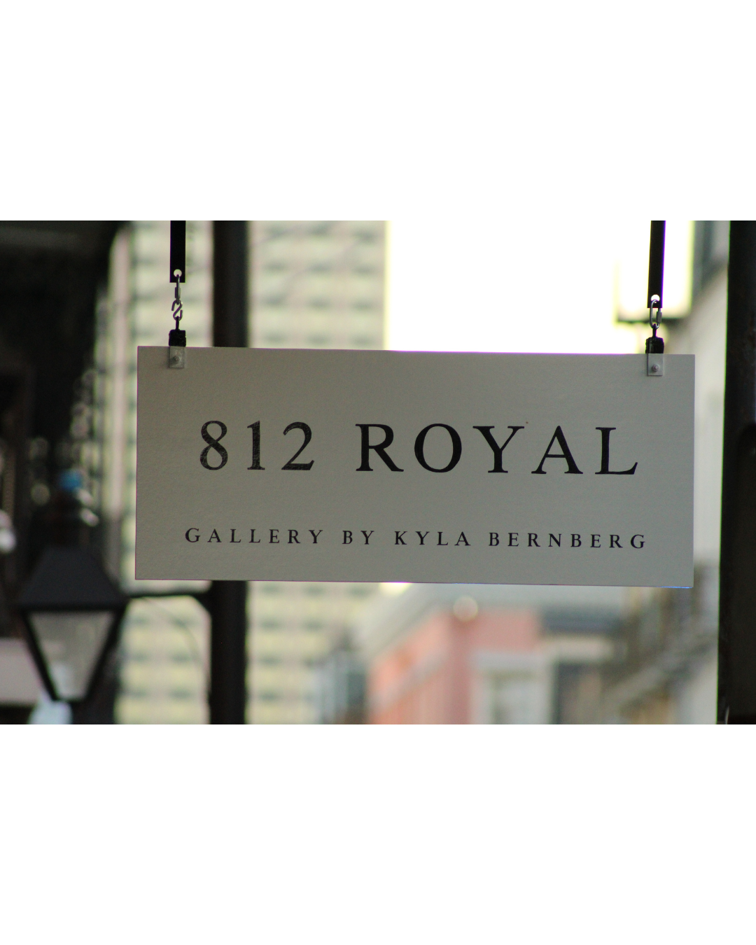 Sign for 812 Royal, Gallery by Kyla Bernberg, hanging on black poles with blurred city buildings in the background.
