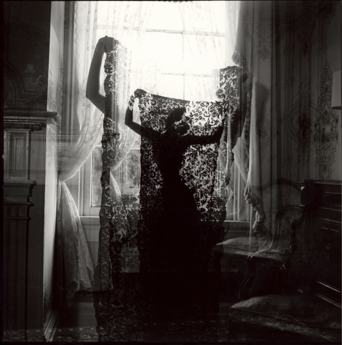 Black and white photo of a person holding patterned curtains in front of a window, with a reflection that creates a layered, artistic effect.