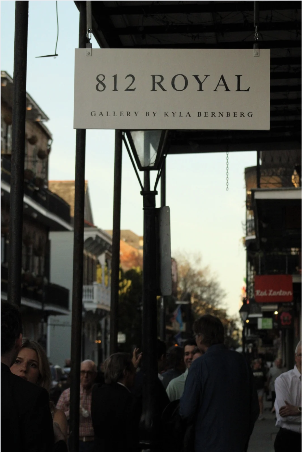 Street sign reading '812 Royal' for an art gallery by Kyla Bernstein, with people standing on a busy sidewalk under the sign in a historic neighborhood, buildings with balconies, and trees in the background.