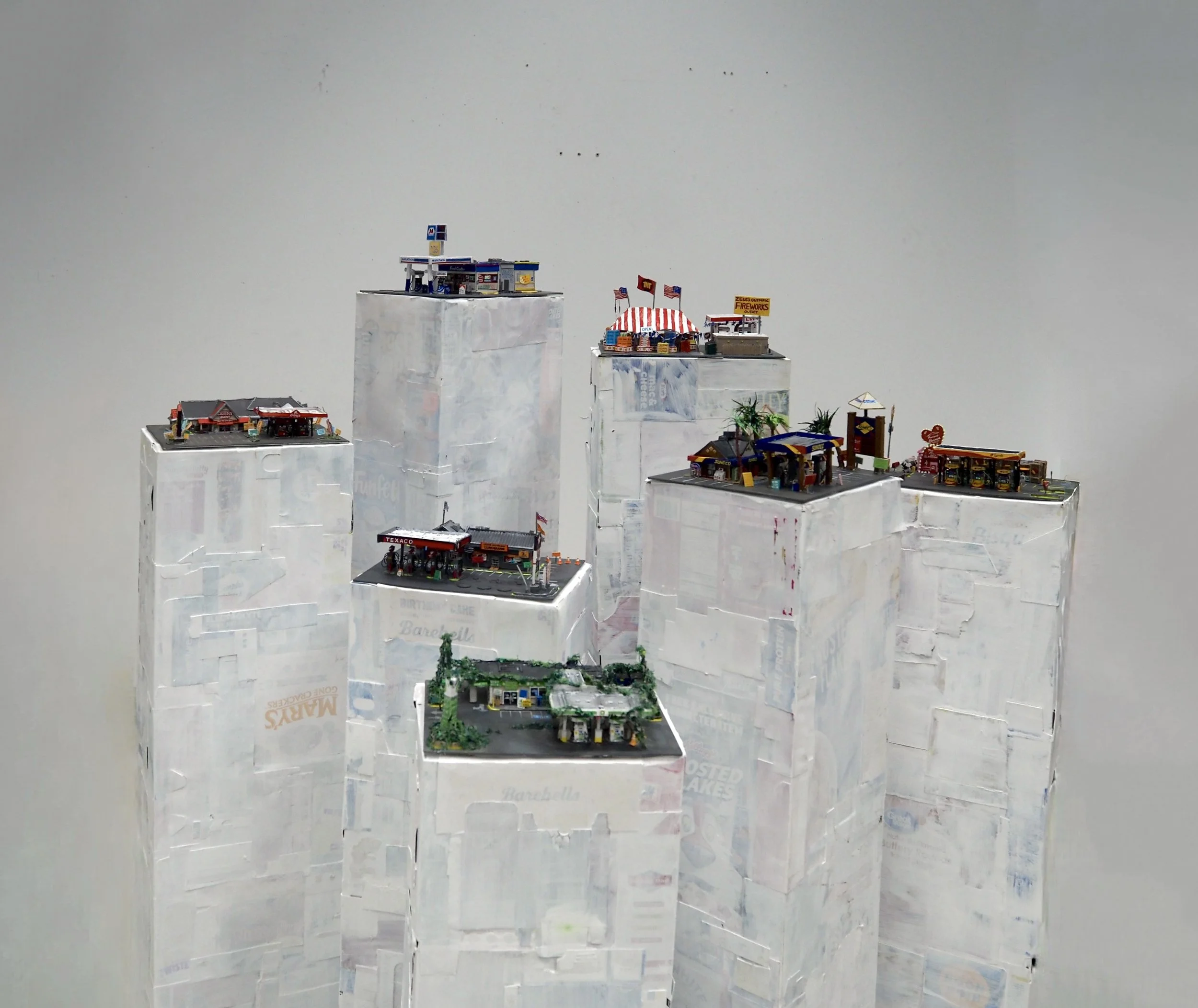 Miniature model buildings resembling a gas station, a storefront with a red roof, and various other small structures, placed on top of white boxes against a plain background.
