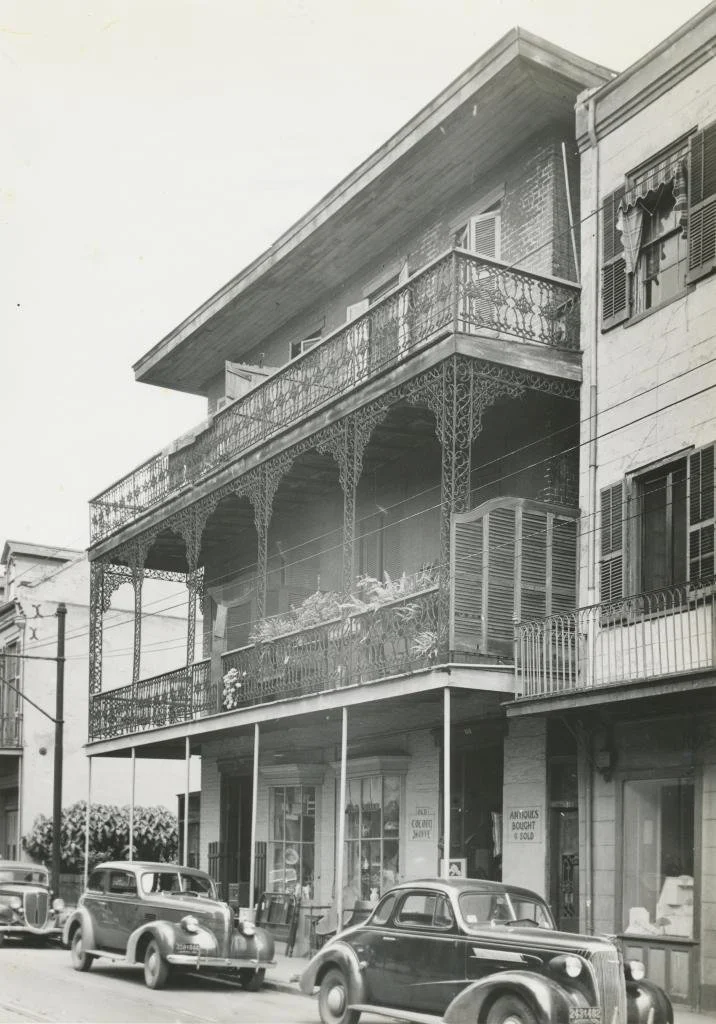 Black and white photo of a three-story building with a balcony on the second floor, decorated with ornate ironwork. The ground floor has shops with large windows and signs, and vintage cars are parked in front.