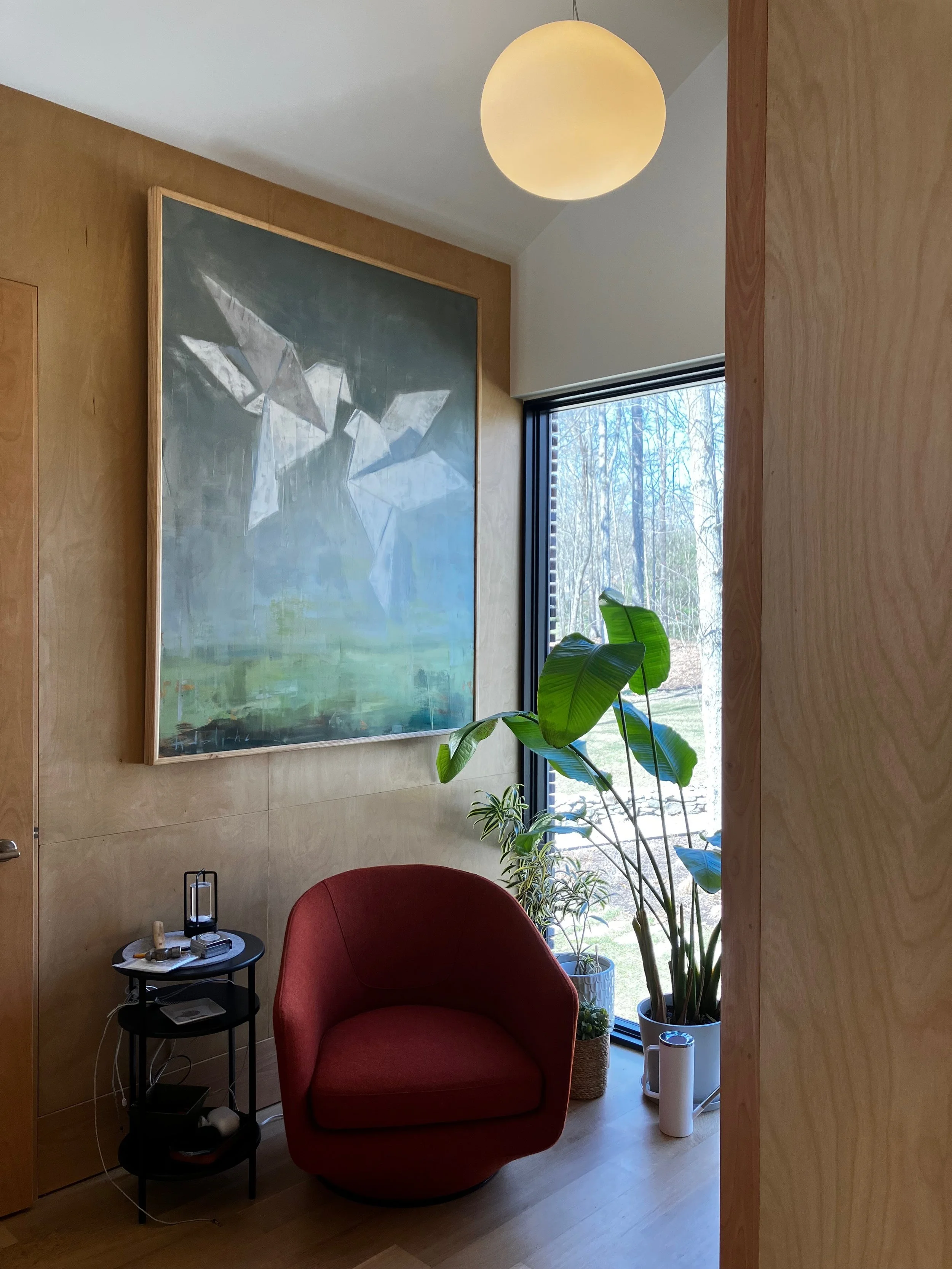 A cozy corner with a red armchair, a black side table holding a small lamp and some items, large potted plants near a window, a painting on a wooden wall, and a round ceiling light, with sunlight and trees outside the window.