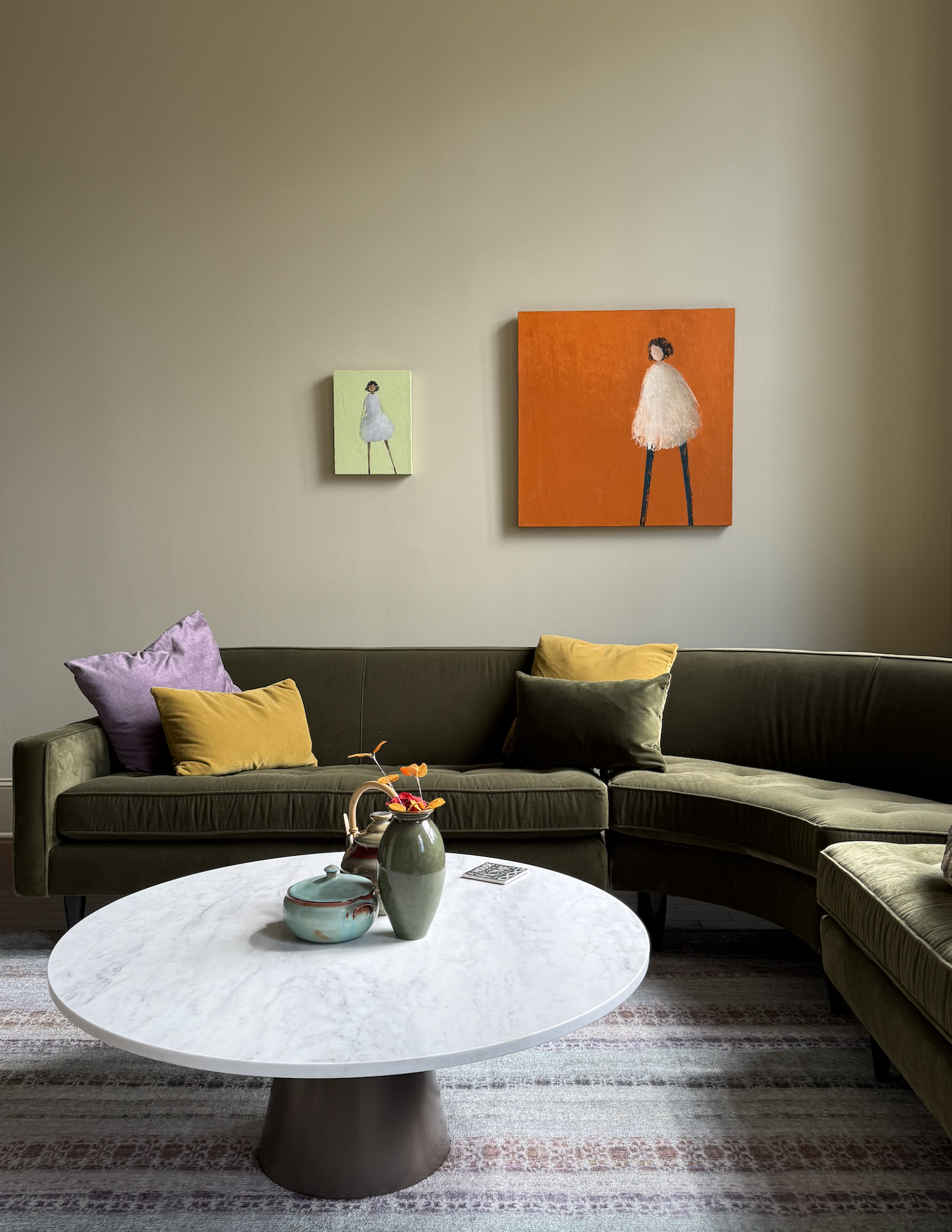 Living room with green sectional sofa, colorful throw pillows, a white marble coffee table with vases, patterned rug, and framed artwork on the wall.
