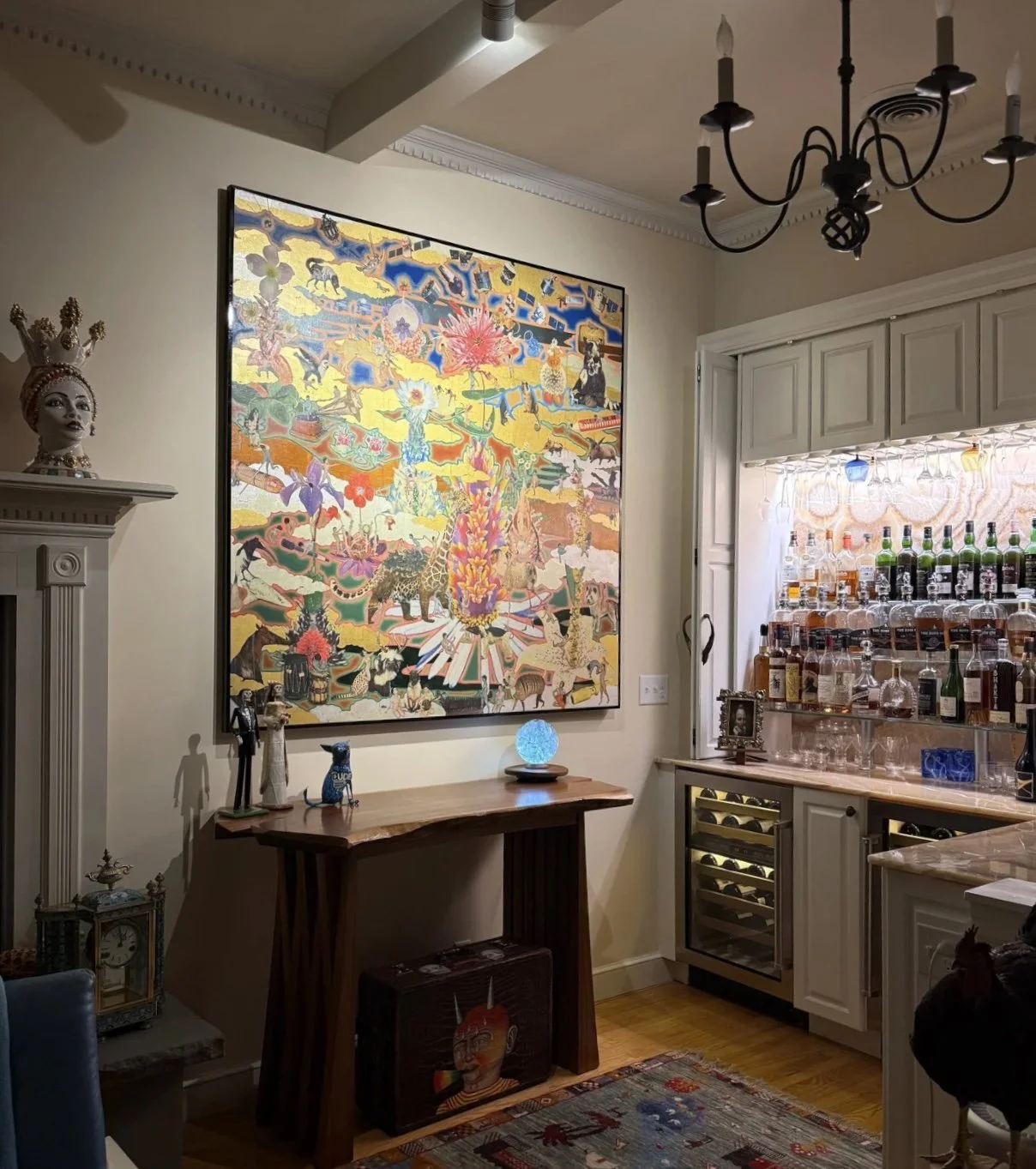 A cozy bar area with a large colorful painting on the wall, various decorative items including a portrait mannequin, a blue crystal ball, and a small framed photo. The bar has bottles of spirits, glassware, and a wine fridge. There is a chandelier hanging from the ceiling and a patterned rug on the wooden floor.