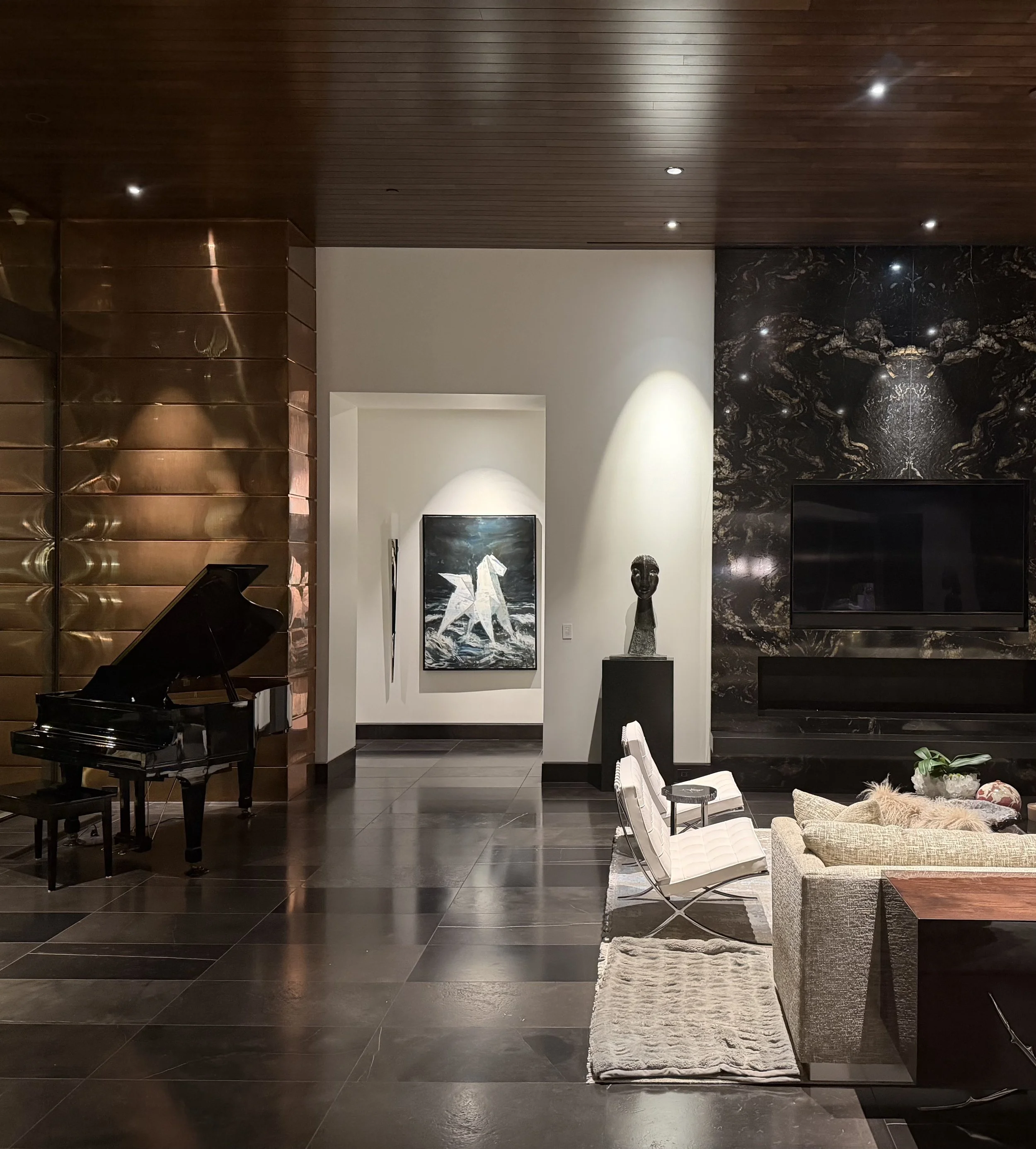 Modern living room with dark wood paneled ceiling, black and brown marble accent wall with a fireplace, a black grand piano, and white leather chairs.