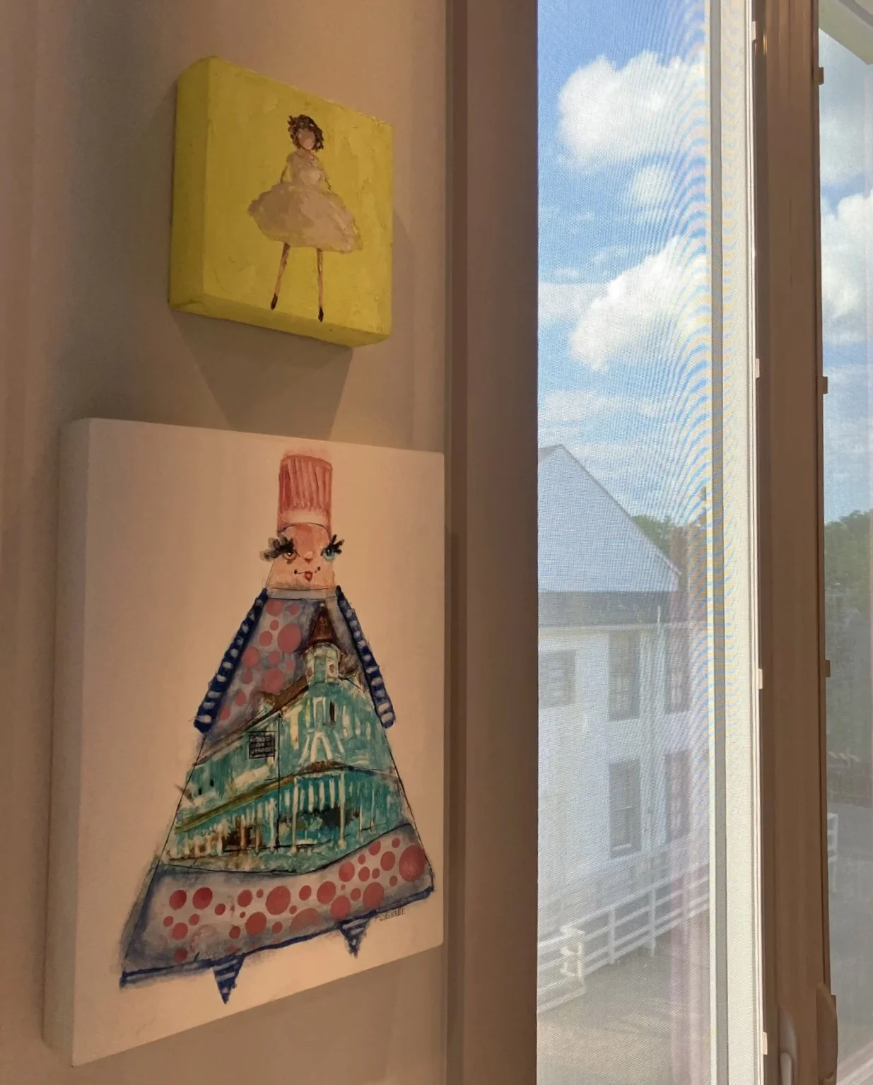 Two pieces of art on a wall near a window: a yellow canvas with a painted girl in a dress and a colorful abstract figure resembling a person with a cityscape on the lower part.