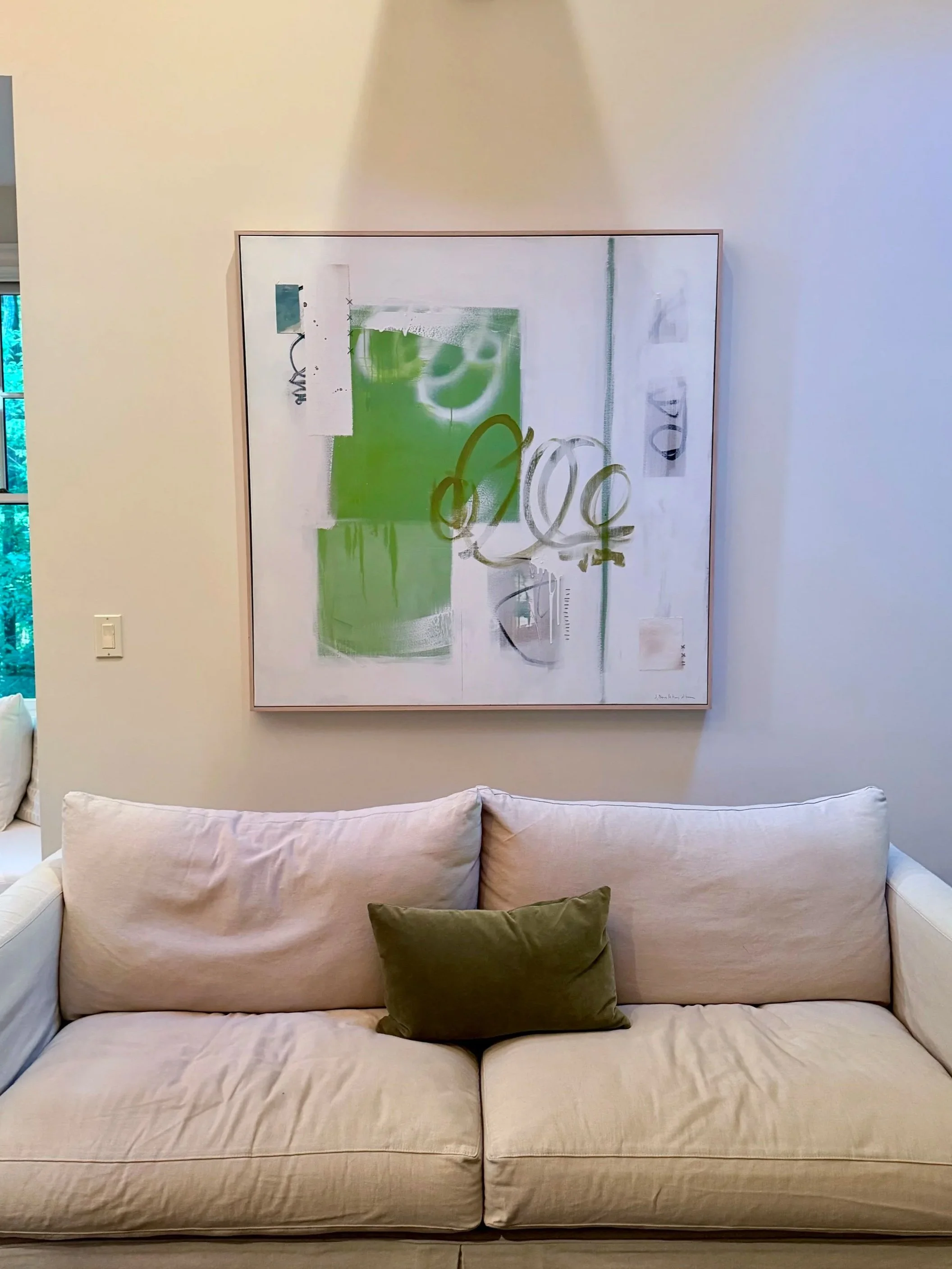 A beige sofa with a small green cushion, in front of a white wall with a large abstract painting above it.