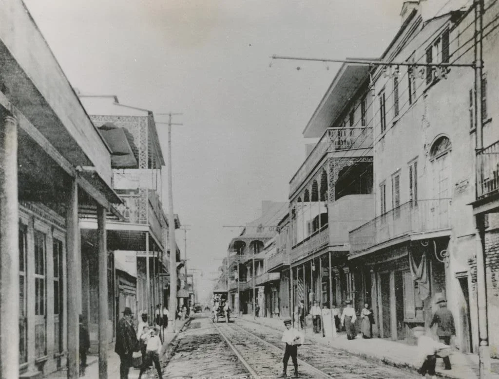 Historical black-and-white photo of a street scene with buildings, streetcars, and people walking on the sidewalks in an early 20th-century city.