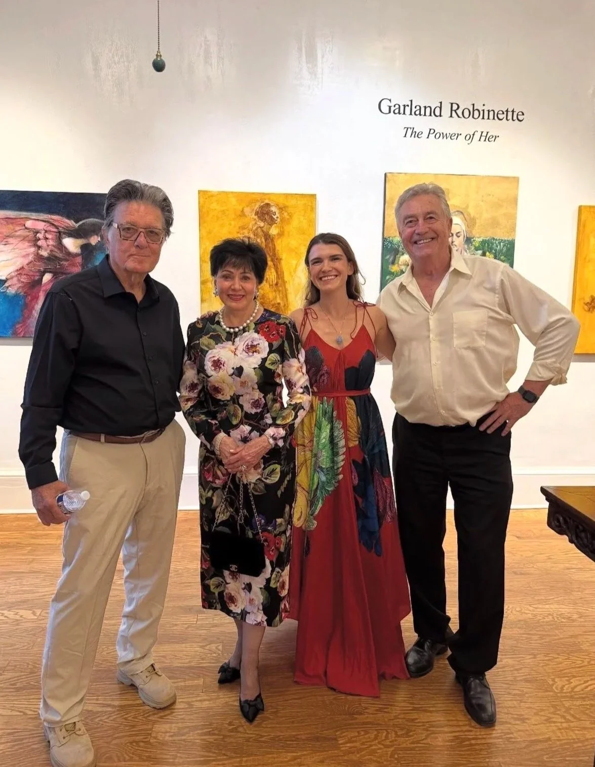 Four people standing in front of a gallery wall with colorful paintings, smiling for a photo, including two men and two women, one woman wearing a floral dress and another woman wearing a vibrant, multicolored dress, with a sign that reads 'Garland Robinette The Power of Her' on the wall.