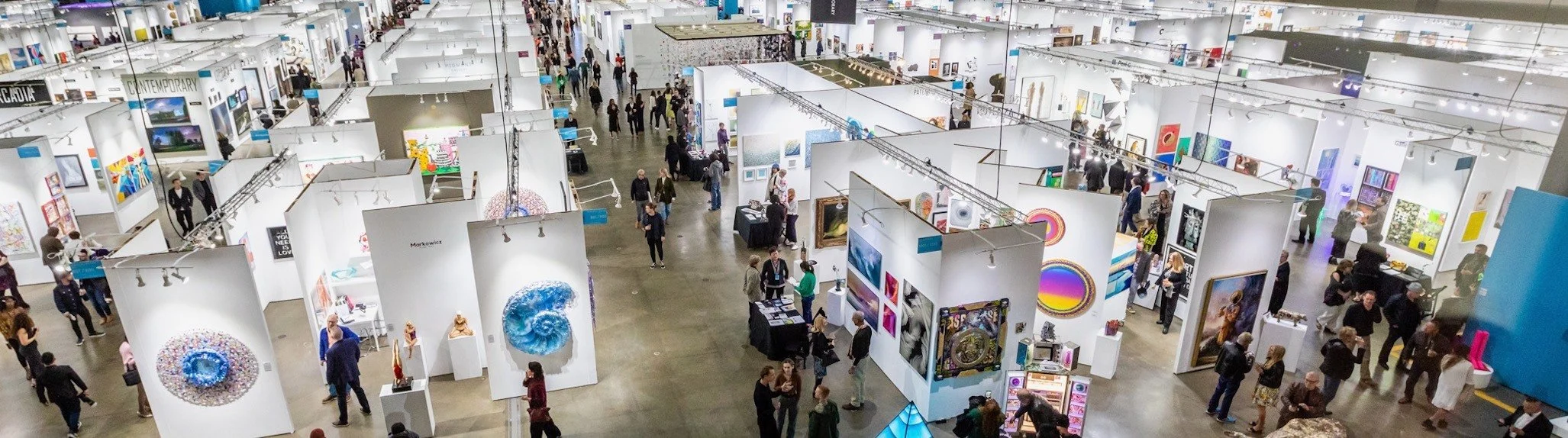 A large art exhibition with numerous white booths displaying various artworks and sculptures. Many visitors are walking around, viewing art pieces, and engaging with artists and other attendees.