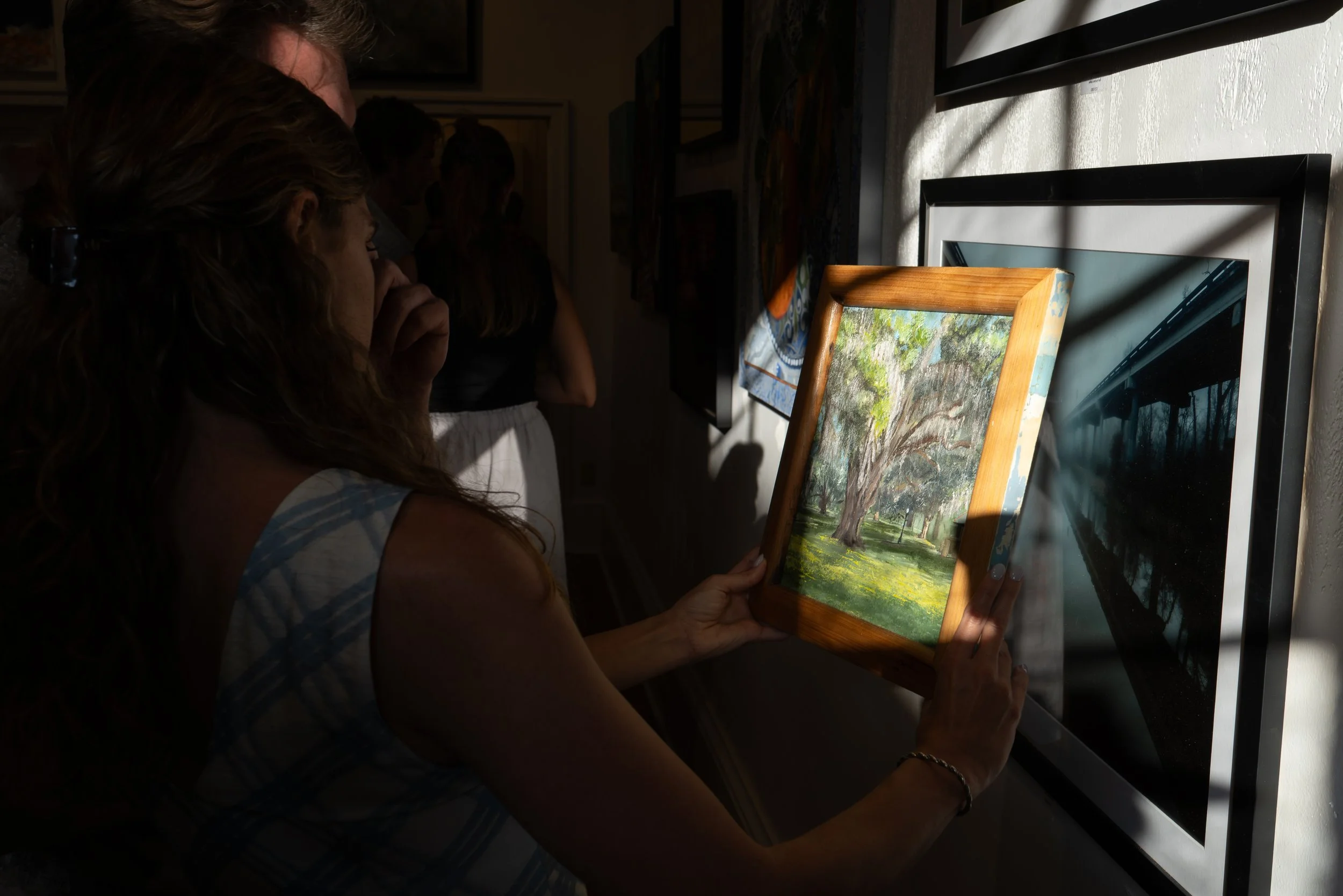 A woman with curly hair holding a framed painting of a park with trees and grass, in an art gallery.