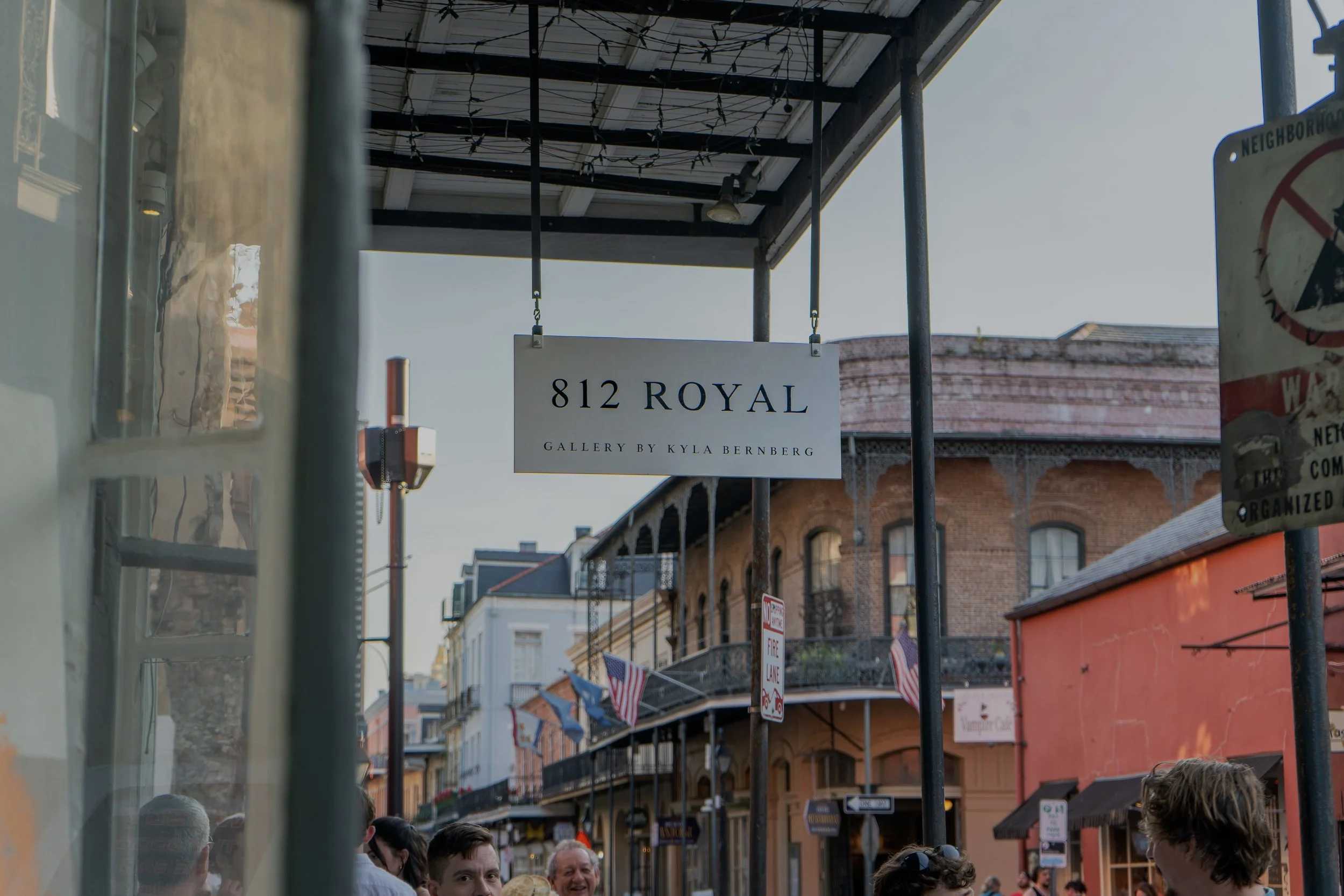 Street scene with a sign reading '812 Royal, Gallery by Kyla Bernberg', people walking, historic brick buildings, storefronts, and American flags hanging across the street.