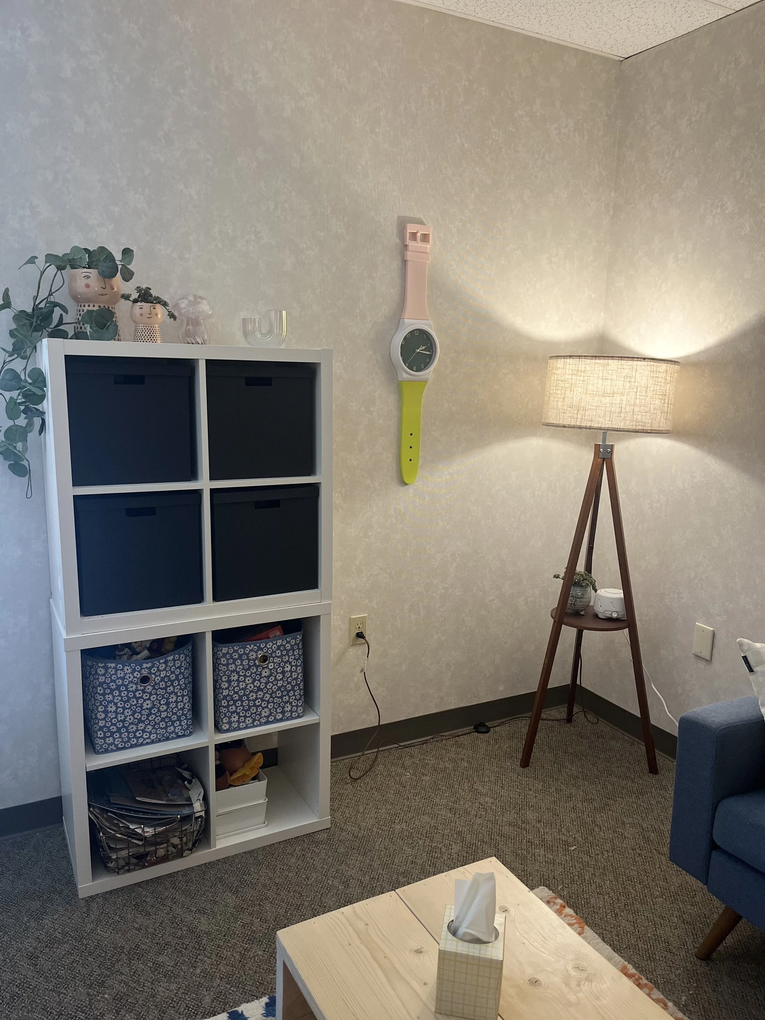 A corner of a living room with a white cube bookshelf, a floor lamp with a beige shade, and a blue sofa. The bookshelf has decorative jars, a plant, and fabric bins. A clock is mounted on the wall, and a tissue box sits on a wooden coffee table in the foreground.