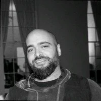 A bearded man with a shaved head smiling in front of a window with curtains.