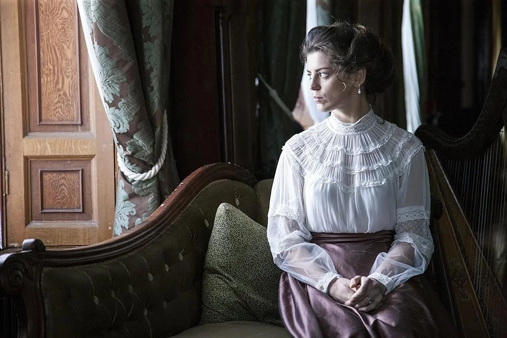 A woman with styled hair and earrings sitting on a vintage dark green tufted sofa near a window with ornate curtains, looking out thoughtfully.