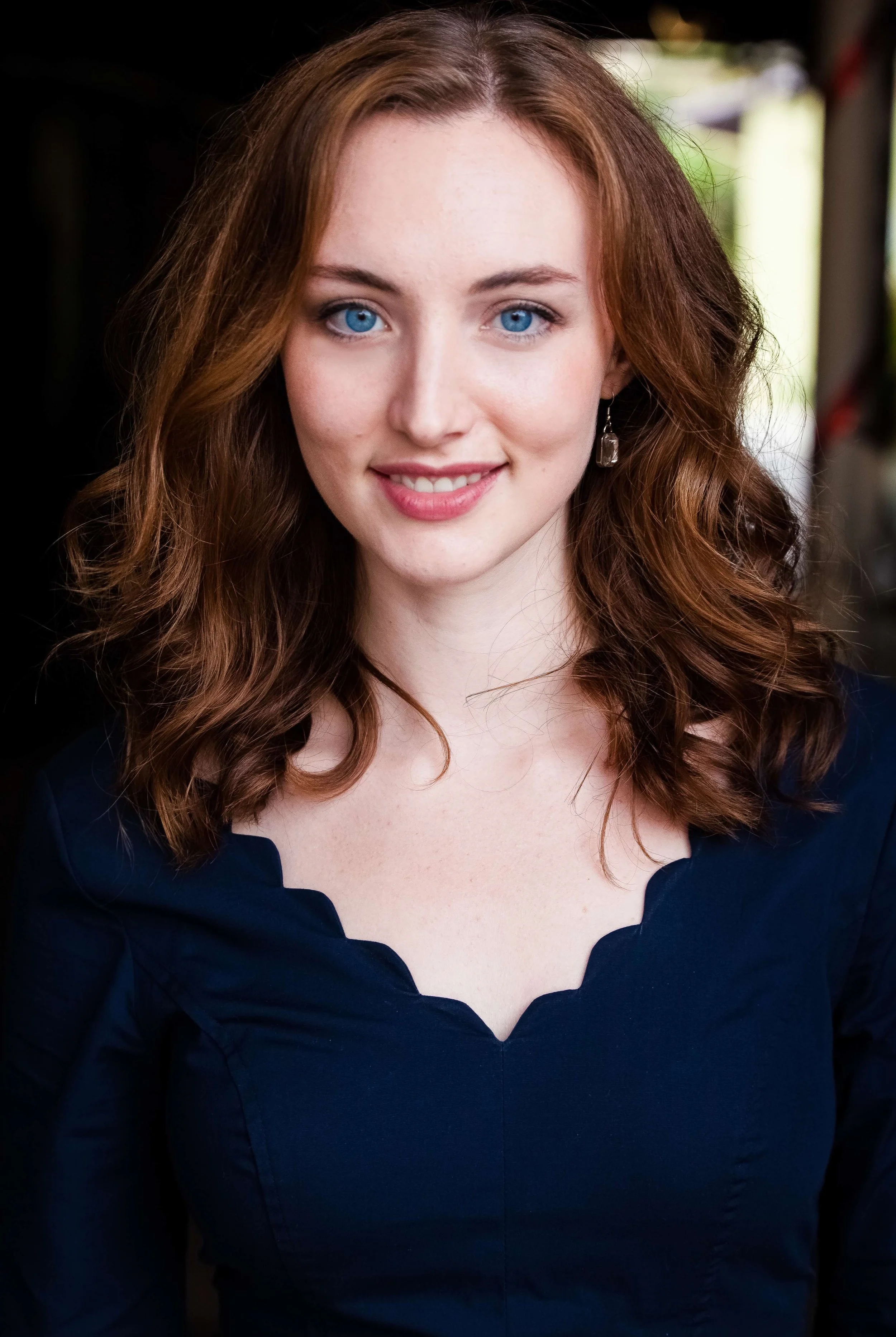 Close-up portrait of a young woman with curly red hair, bright blue eyes, and fair skin, wearing a navy blue top and earrings, smiling softly.