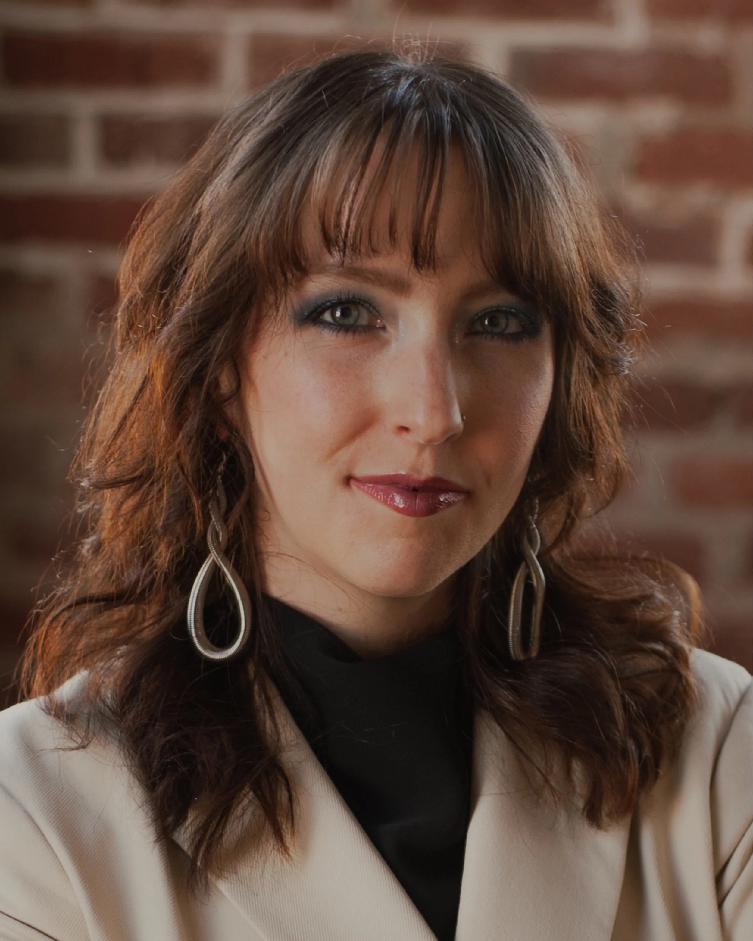 Close-up of a woman with brown hair, grey-green eyes, wearing silver hoop earrings, a black top, and a beige blazer, against a brick wall background.
