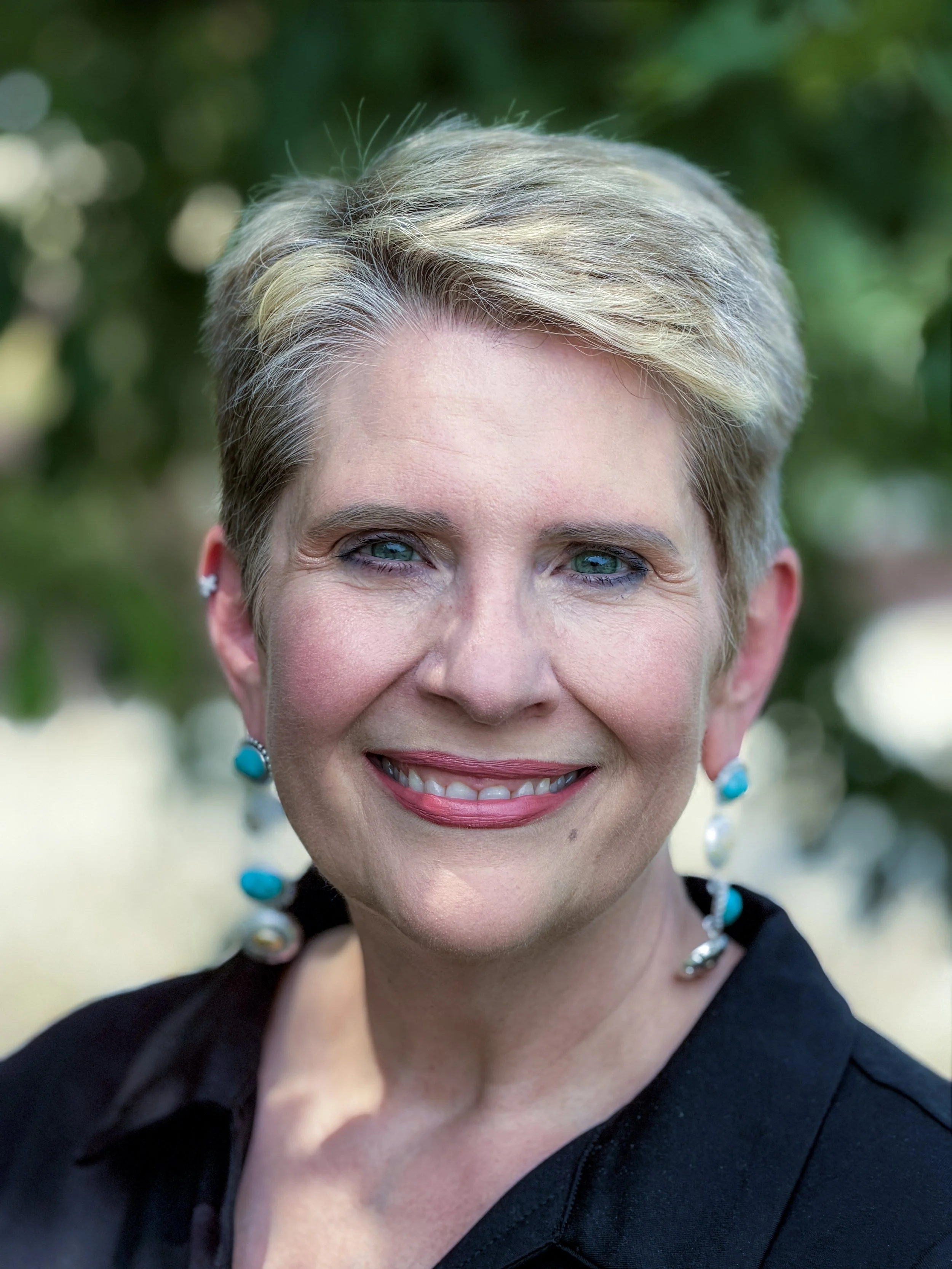 Close-up of a smiling woman with short, blonde hair, wearing earrings and a black top, outdoors with a blurred green background.