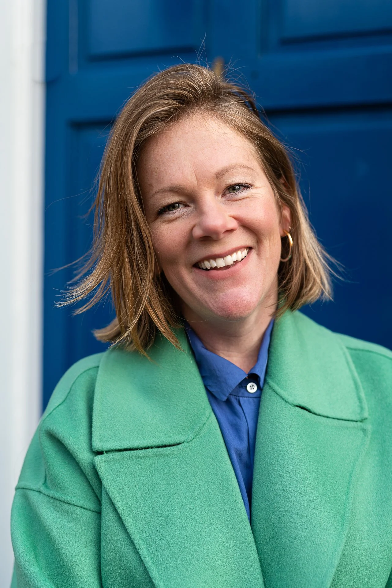 A photograph of Hannah Reichardt, with shoulder-length light brown hair smiling, wearing a bright green coat and a blue shirt, standing in front of a blue door.