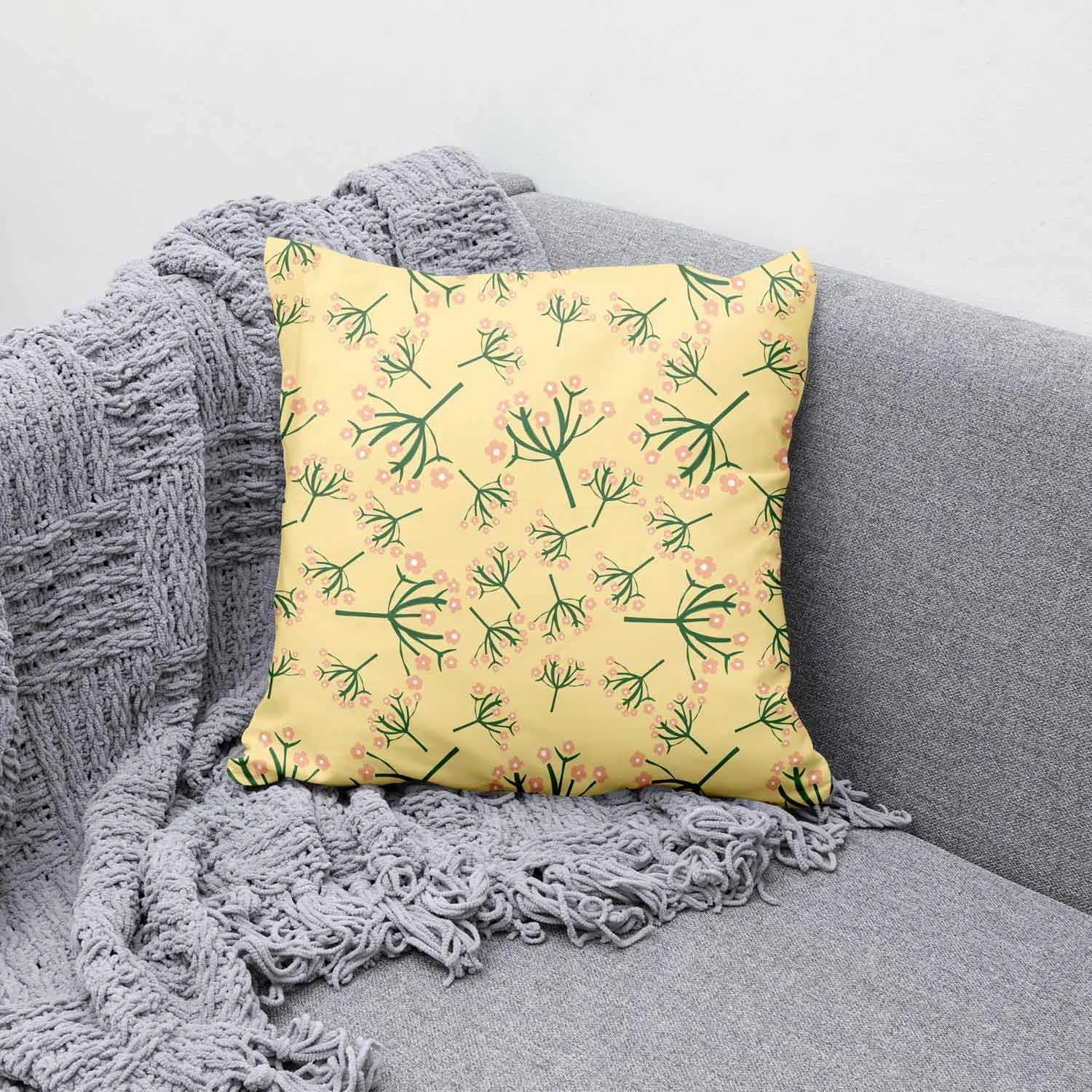 A decorative pillow with a yellow background and a green and pink floral pattern resting on a gray upholstered sofa, partially covered by a chunky knit gray blanket.