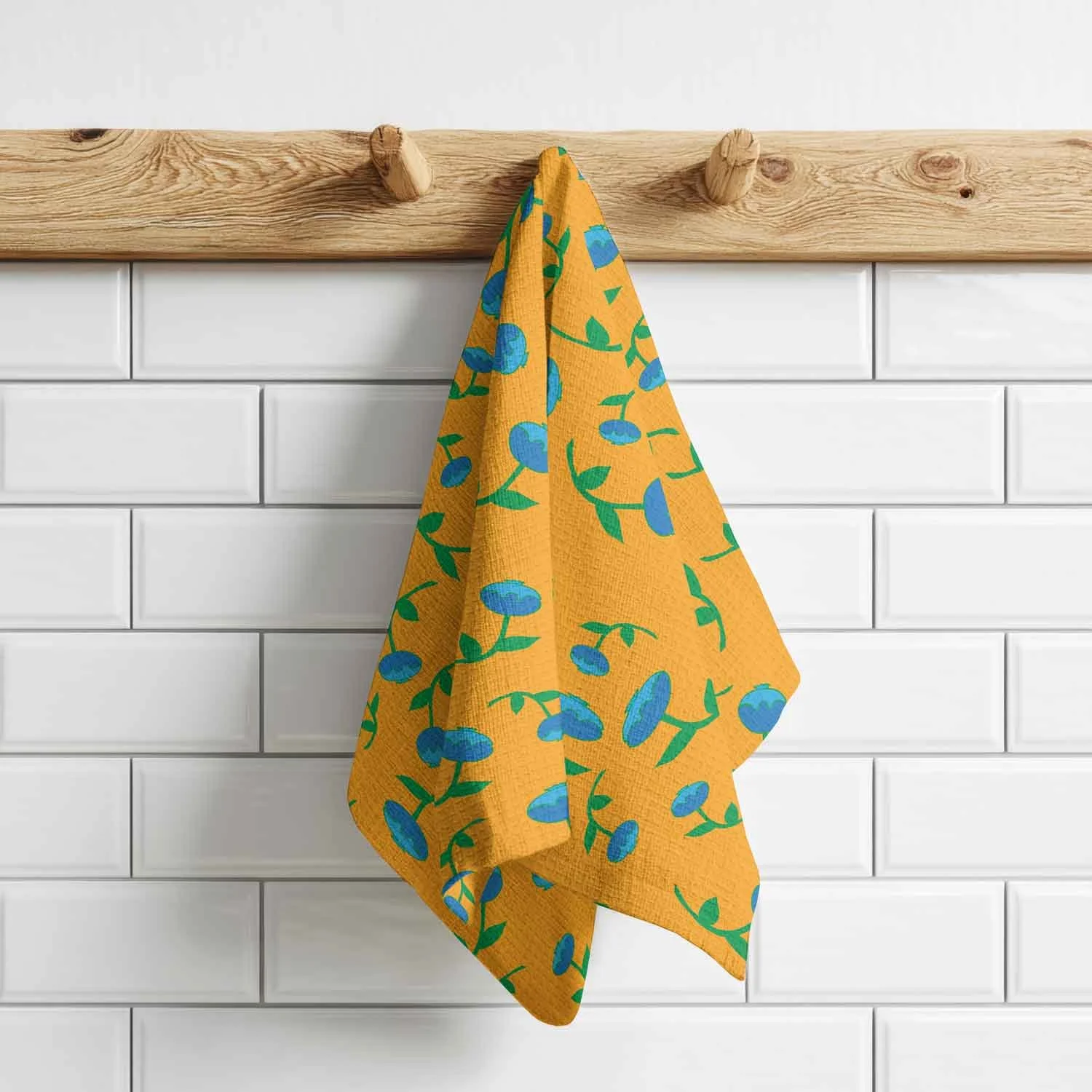 A yellow kitchen towel with a blueberry pattern hanging on a wooden hook against a white subway-tiled wall.