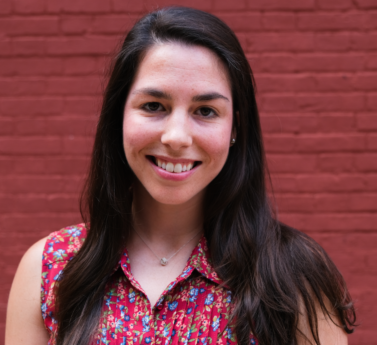 Portrait of a young woman with long dark hair, smiling, standing in front of a red brick wall, wearing a colorful floral sleeveless top and a silver necklace.
