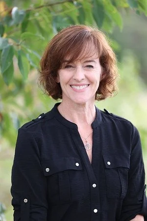 A woman with short, wavy brown hair smiling outdoors with green foliage in the background.