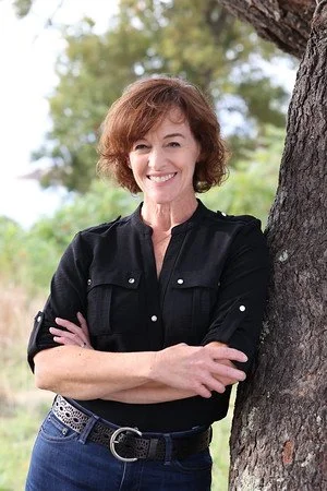 A woman with short brown hair smiling outdoors, standing next to a tree with greenery in the background.