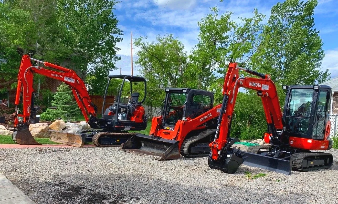 Three mini excavators parked on a gravel surface with green trees and a blue sky in the background.