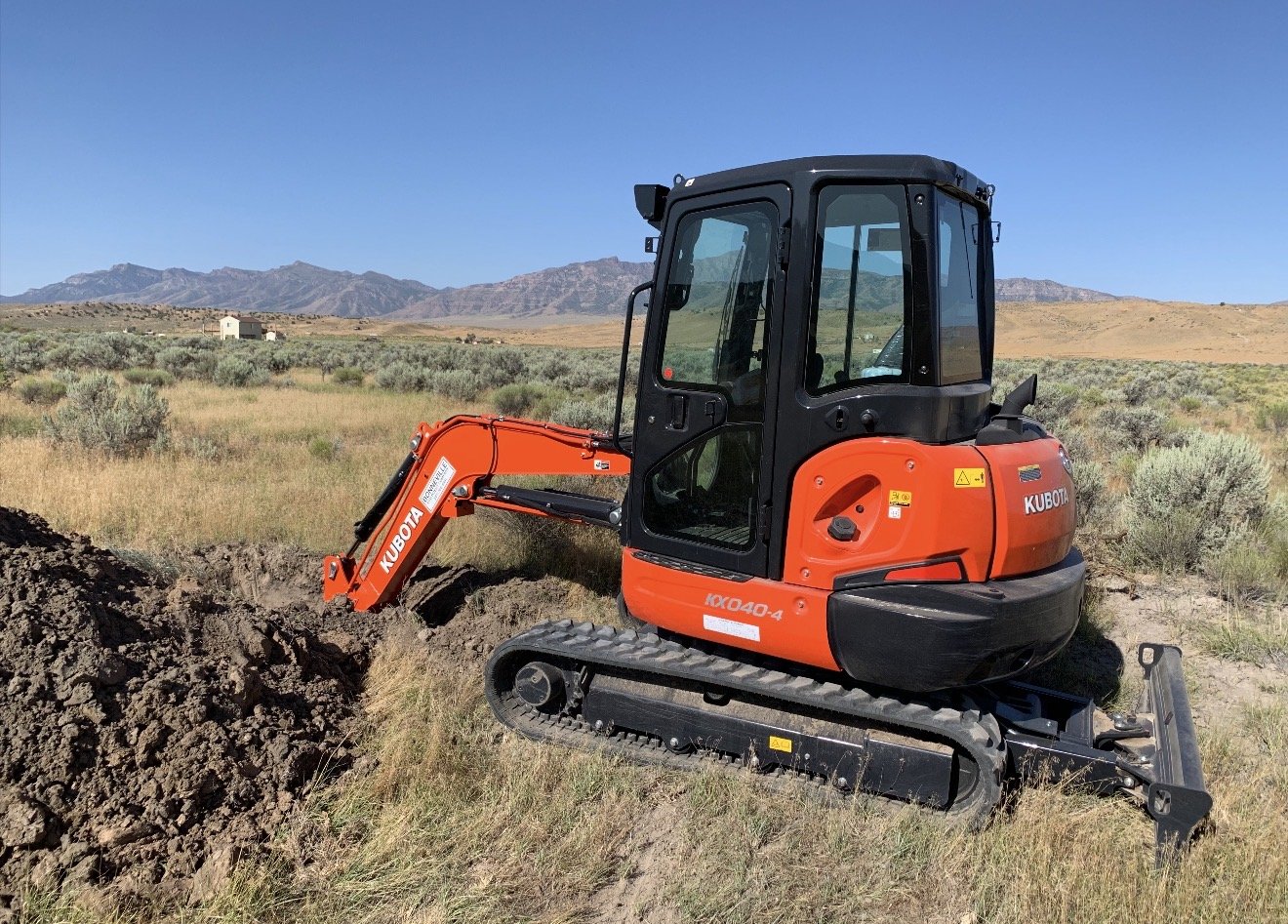 Orange Kubota mini excavator digging into the ground in a grassy field with mountains in the background under a clear blue sky.
