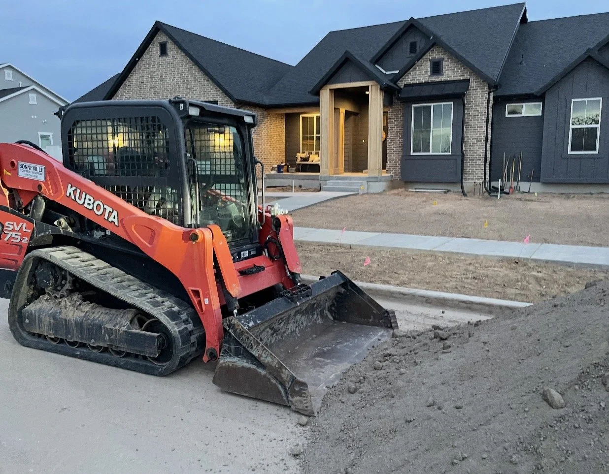 Construction site in front of a house with a trench being dug by a red skid steer loader, with a sidewalk and house exterior with front porch visible in the background.