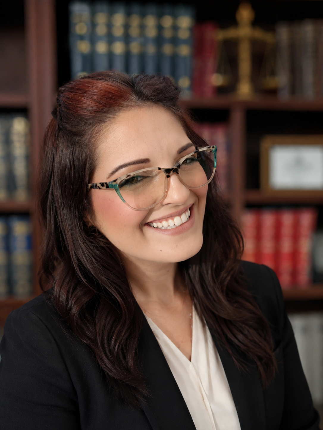 A woman with long dark wavy hair, glasses, and a white blouse, smiling in front of a bookshelf filled with books and a framed photo.