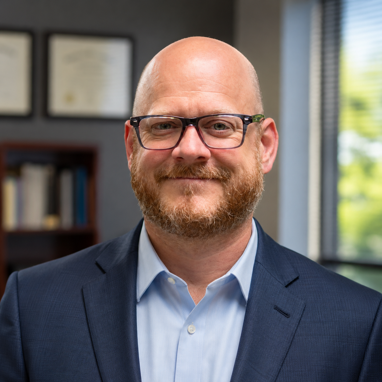 A smiling man with a beard wearing glasses, a navy suit, and a light blue shirt. He is indoors in an office.