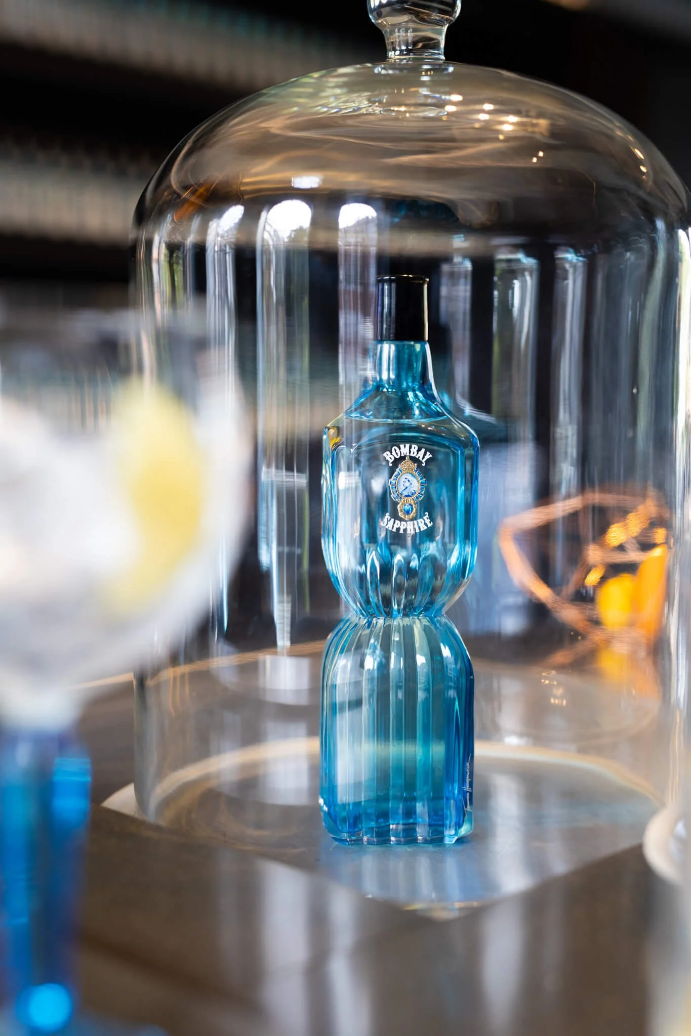 A glass cloche covering a blue Bombay Sapphire gin bottle, with reflections of light on the glass surface.