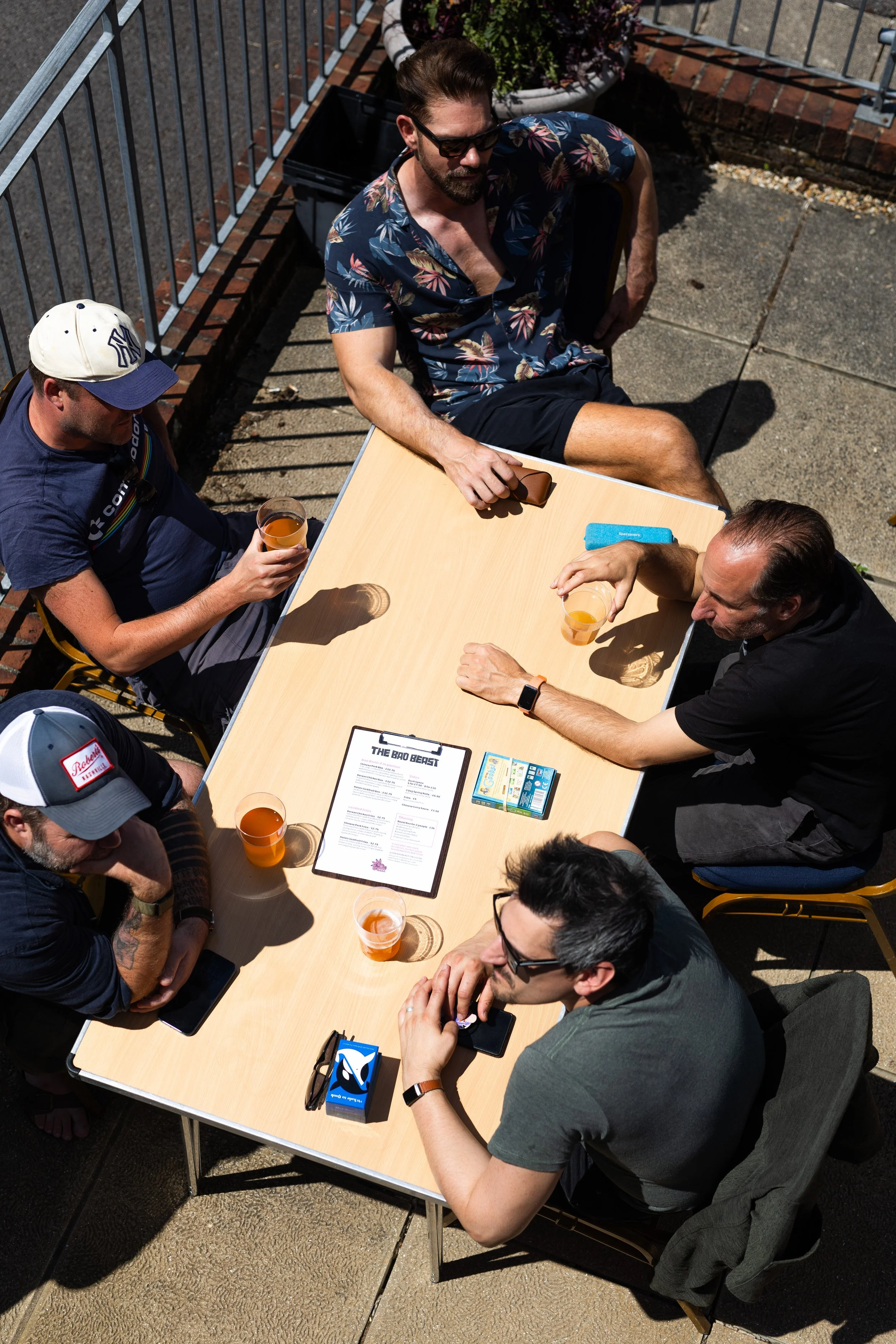 A group of people gathered around an outdoor table, socializing and drinking beverages on a sunny day. There are six visible individuals, some holding drinks and using their phones. The table has a menu and some small boxes on it.