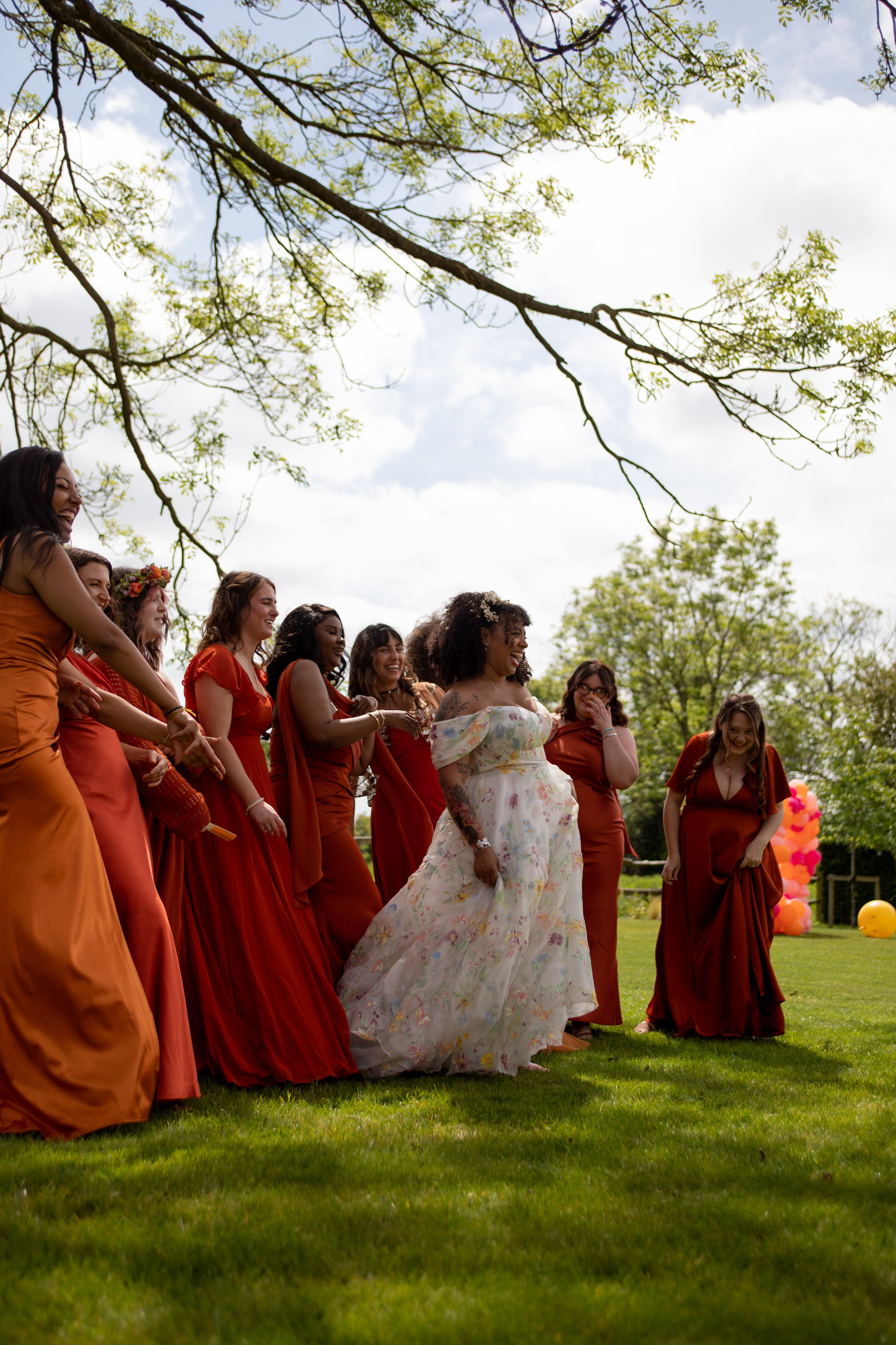 A group of women dressed in matching burnt orange and rust-colored dresses standing outdoors on a grassy field, with some women smiling and laughing. One woman in the center wears a white floral dress, and there are colorful balloons in the backgroun