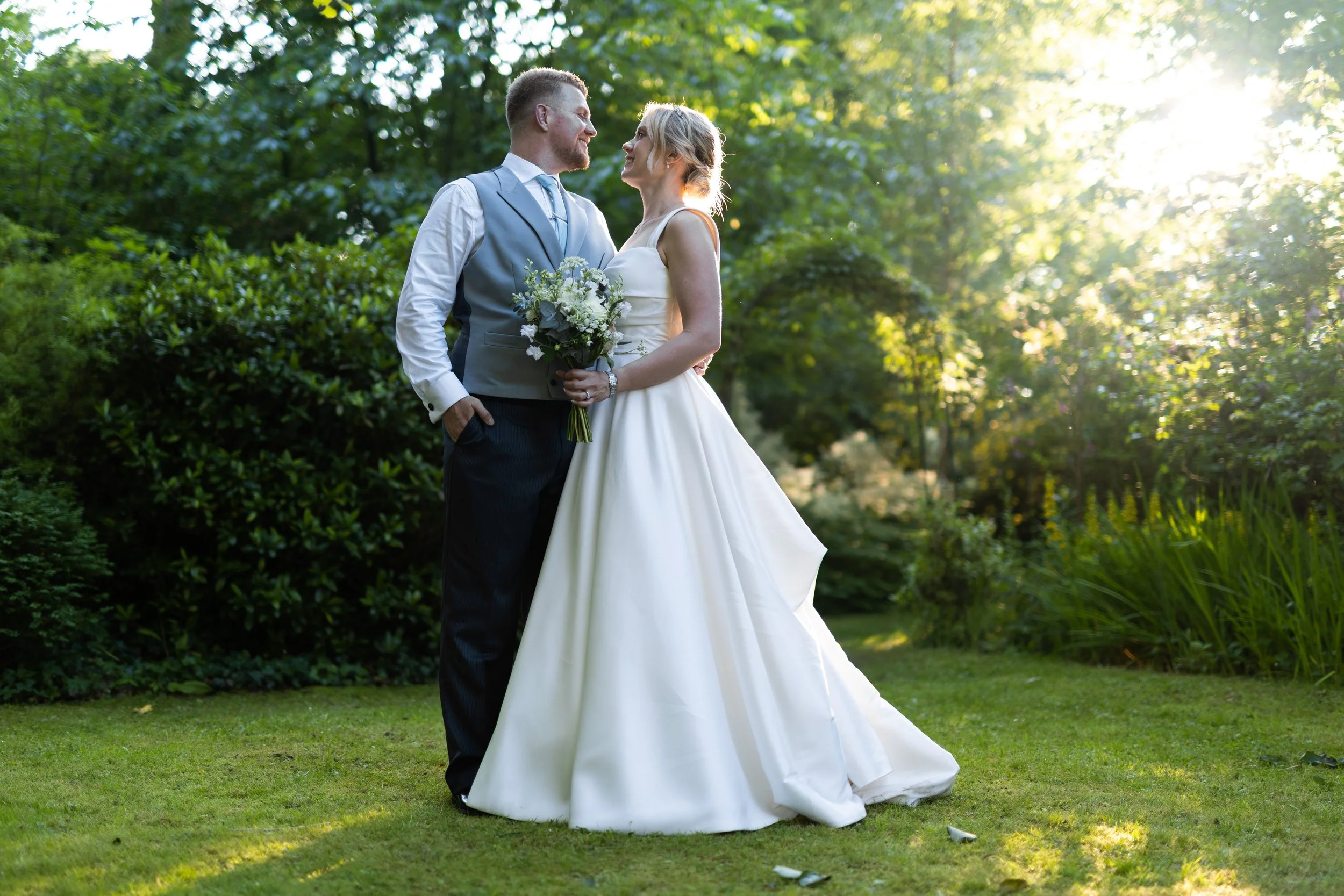 A couple in wedding attire standing close together outdoors, smiling and looking into each other's eyes with a lush green background and sunlight filtering through trees.