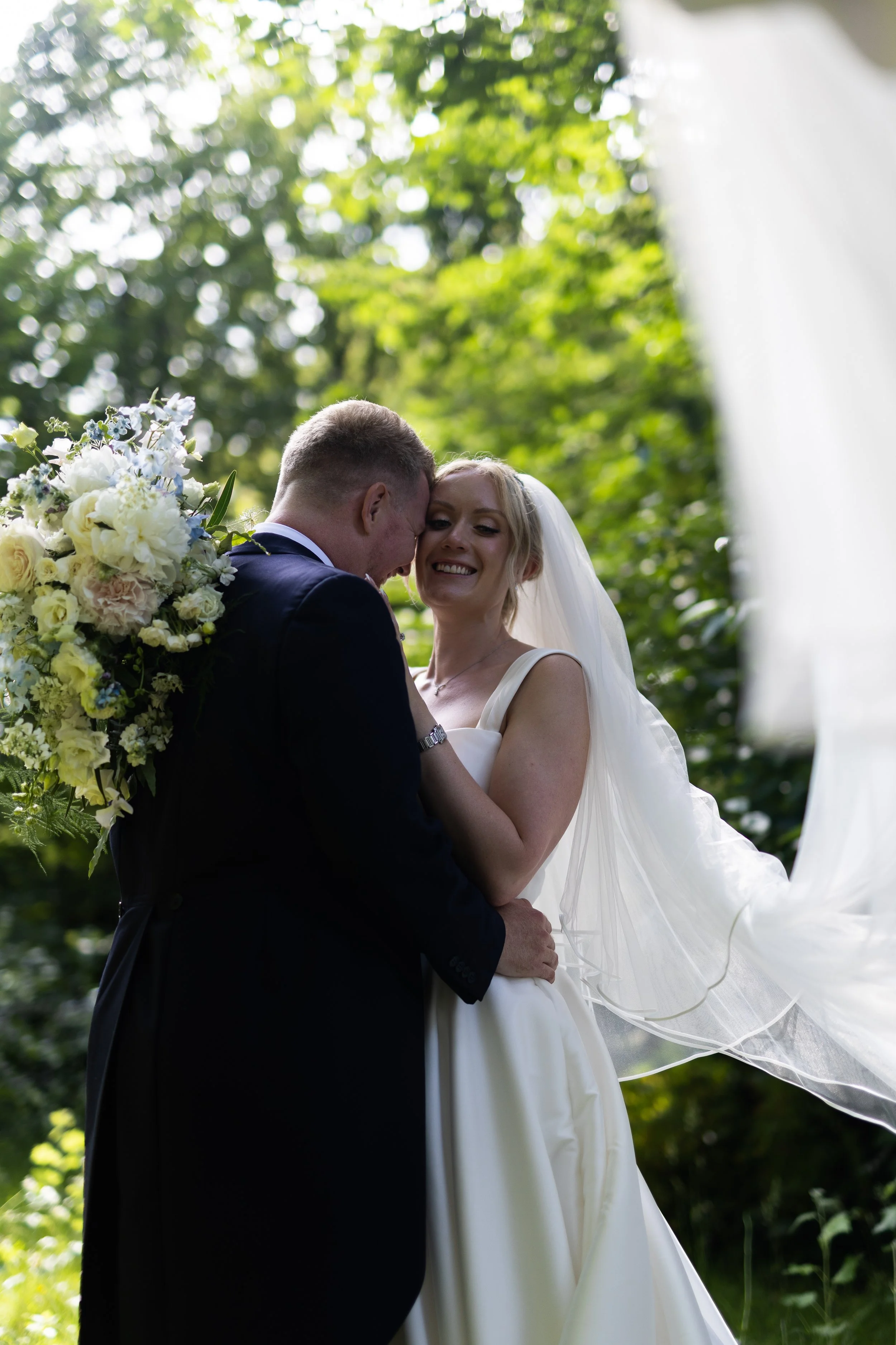 A bride and groom sharing a moment outdoors, with the bride smiling and the groom leaning in close, surrounded by lush green trees. The bride is wearing a white wedding dress and veil, and the groom is dressed in a dark suit. The bride holds a bouque