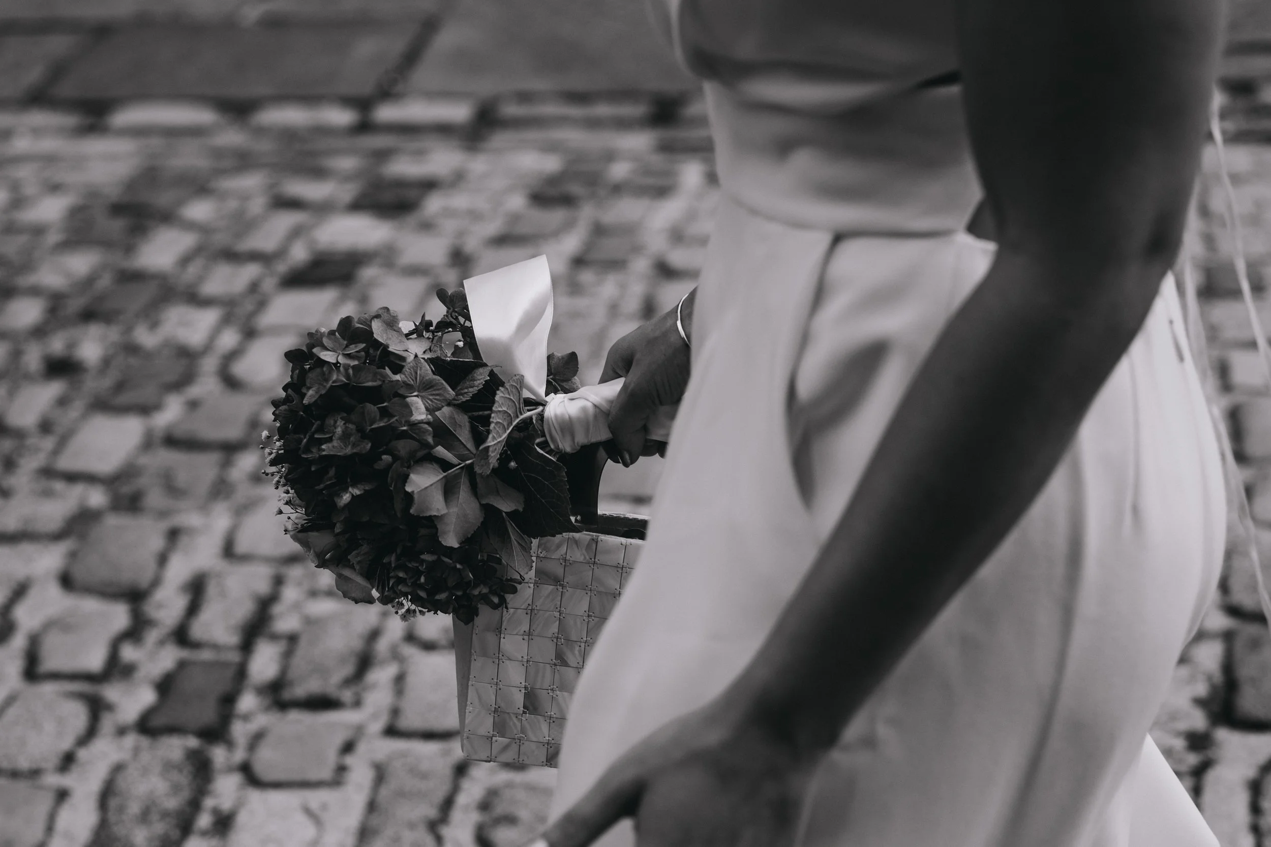 A person dressed in white holding a bouquet of flowers, viewed from behind, with cobblestone street in the background.