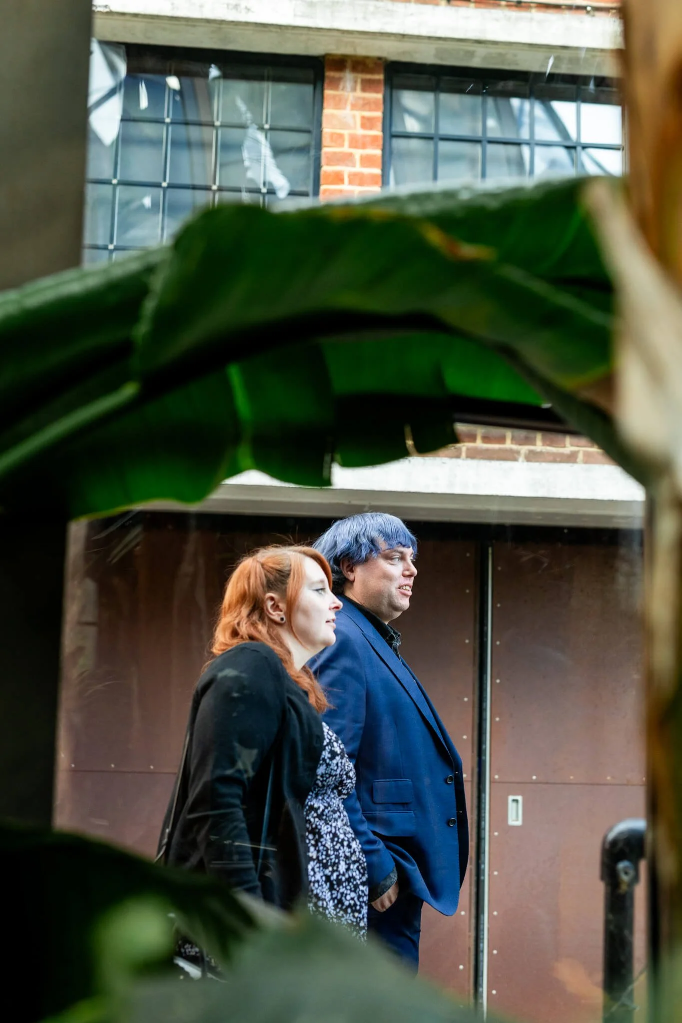 Two people, a woman with red hair and a man with blue hair, standing side by side against a wall. The photo is taken through a gap with green leaves partially obscuring the view.