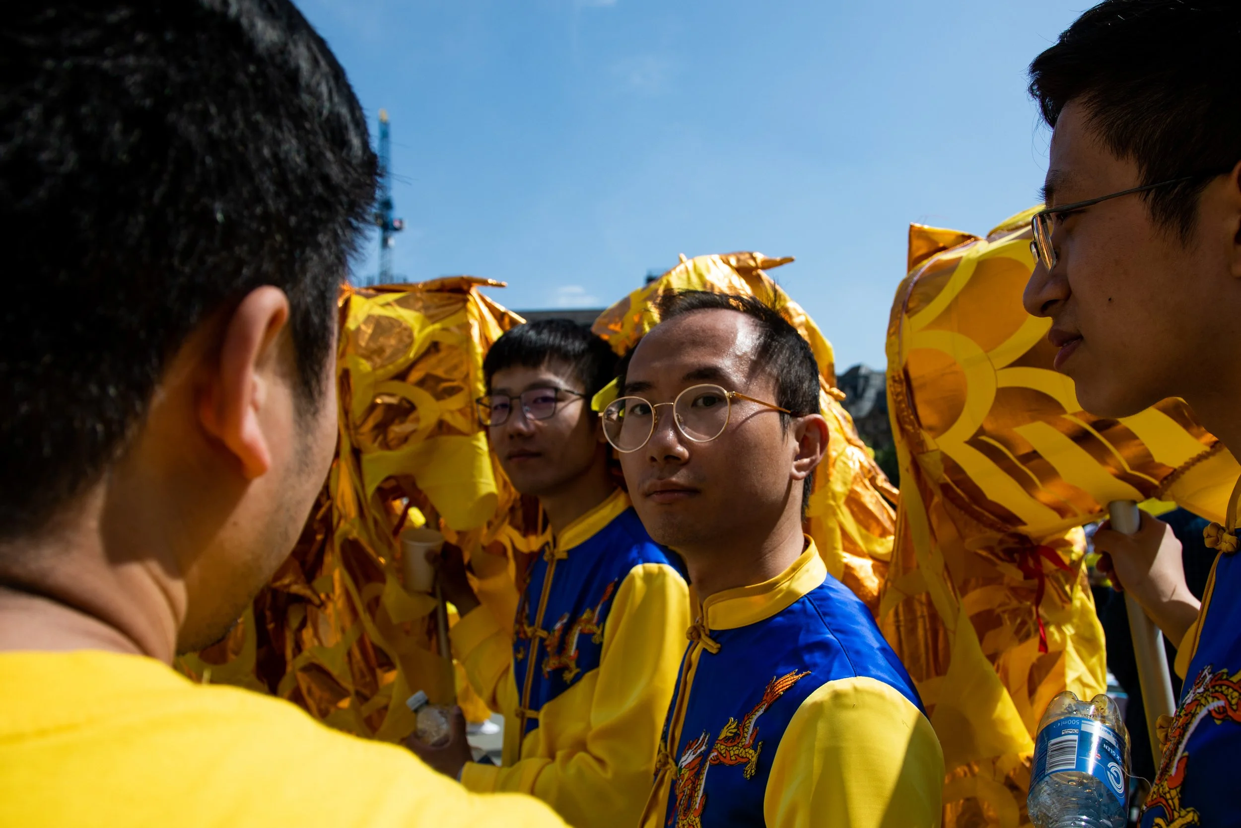 A group of people wearing yellow and blue Asian traditional costumes with dragon designs, standing in a circle, outdoors under a clear blue sky, engaged in a discussion or activity.