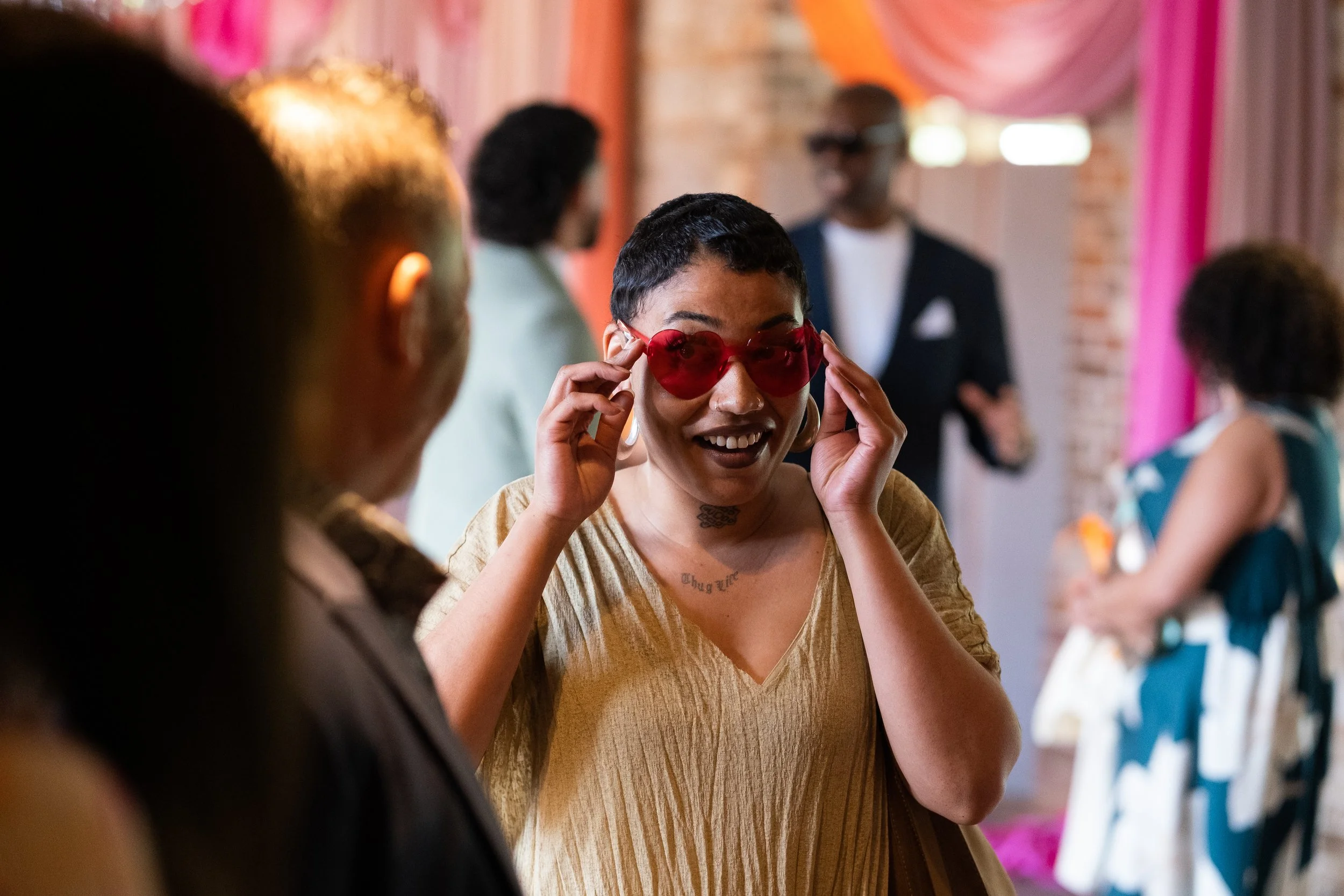 A woman with short dark hair and tattoos on her neck is adjusting red heart-shaped sunglasses and smiling at an indoor party or event with colorful decorations and other people in the background.