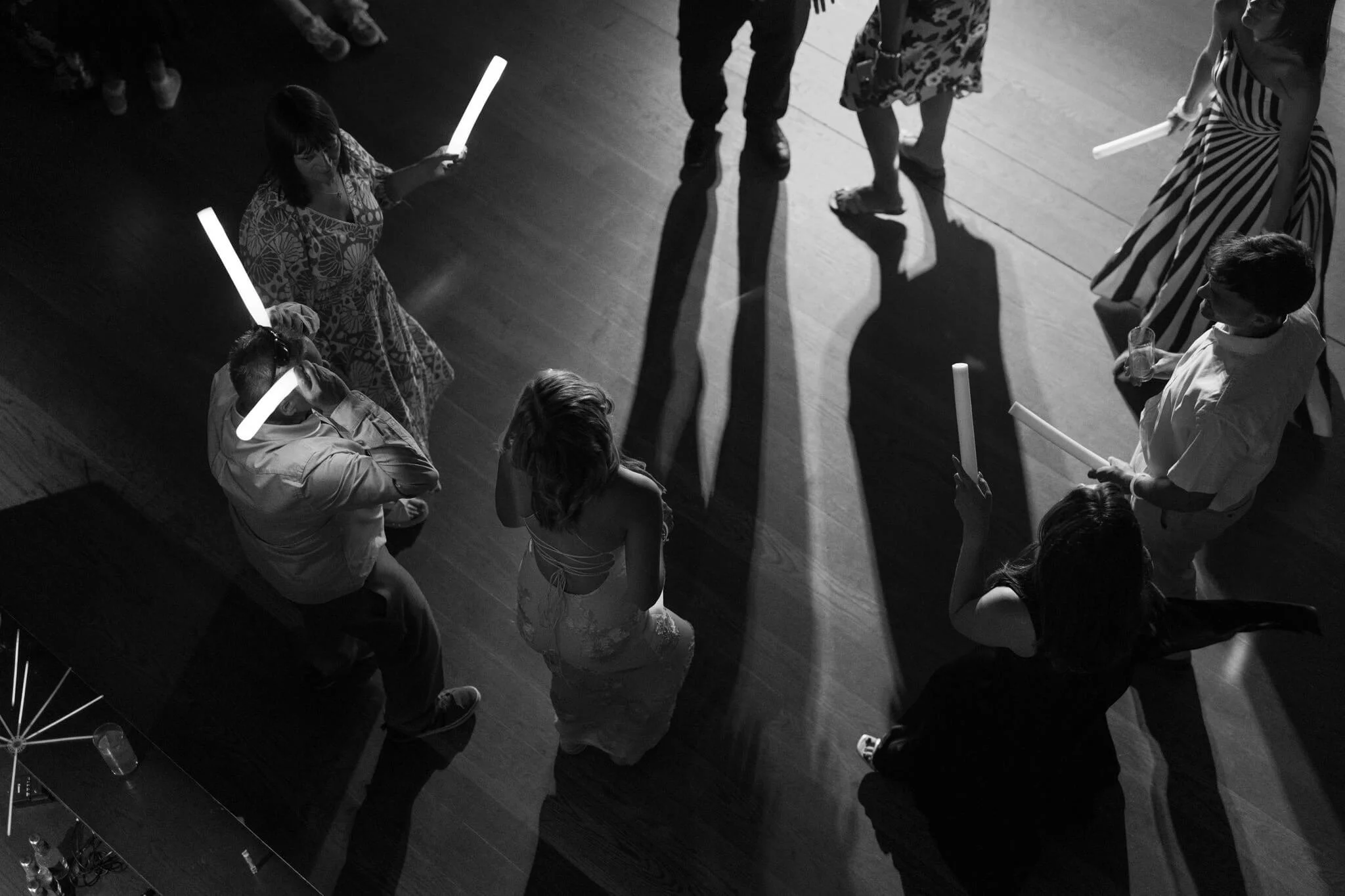 People gathered in a circle holding glow sticks, with their shadows cast on a wooden floor, suggesting a dance or celebration event.