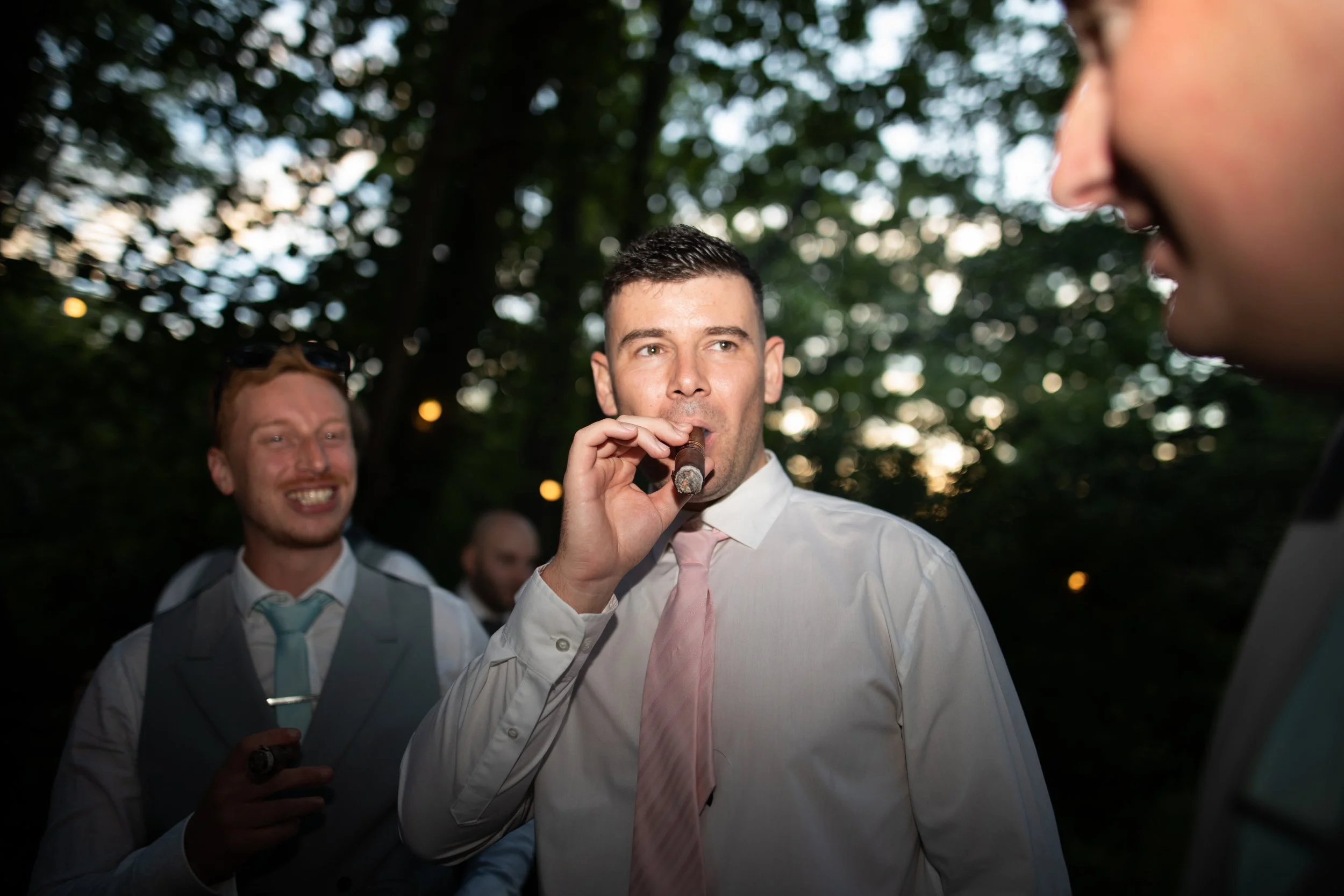 A man in a white shirt and pink tie holding a cigar, standing outdoors with other people around him at dusk, with trees in the background.