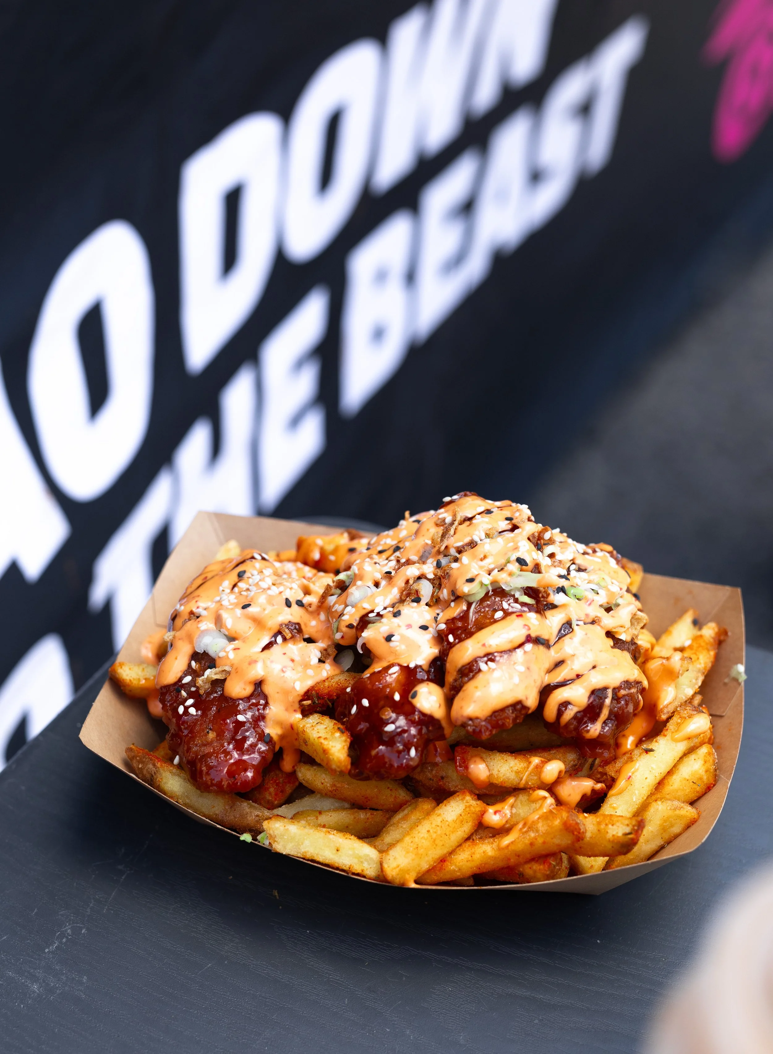 A serving of loaded fries topped with cheese, sauce, and herbs on a paper tray, with a black background that has the words '#Doughbox' in large white letters.