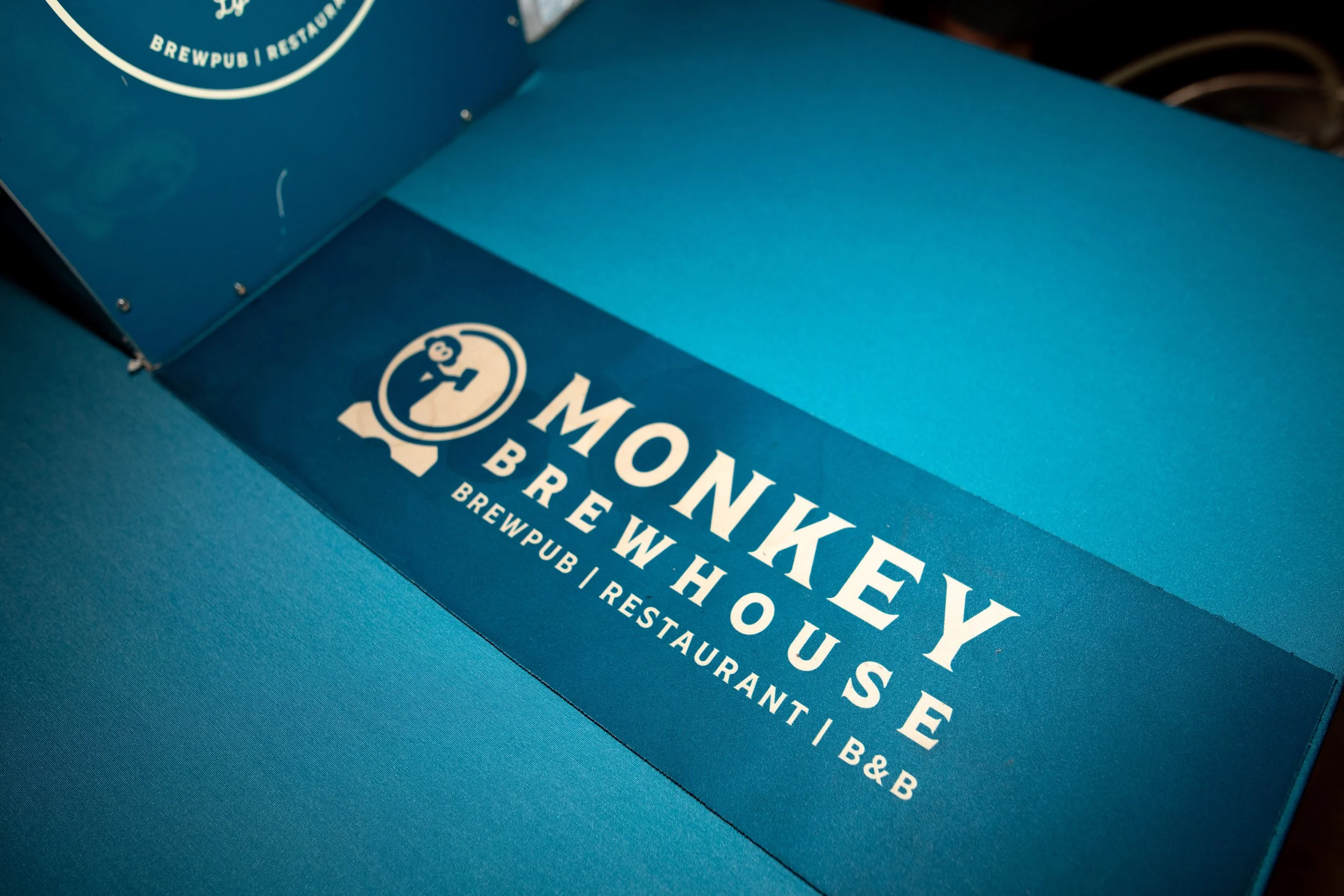 Blue takeout box from Monkey Wrench Brewery with white text, placed on a dark surface.