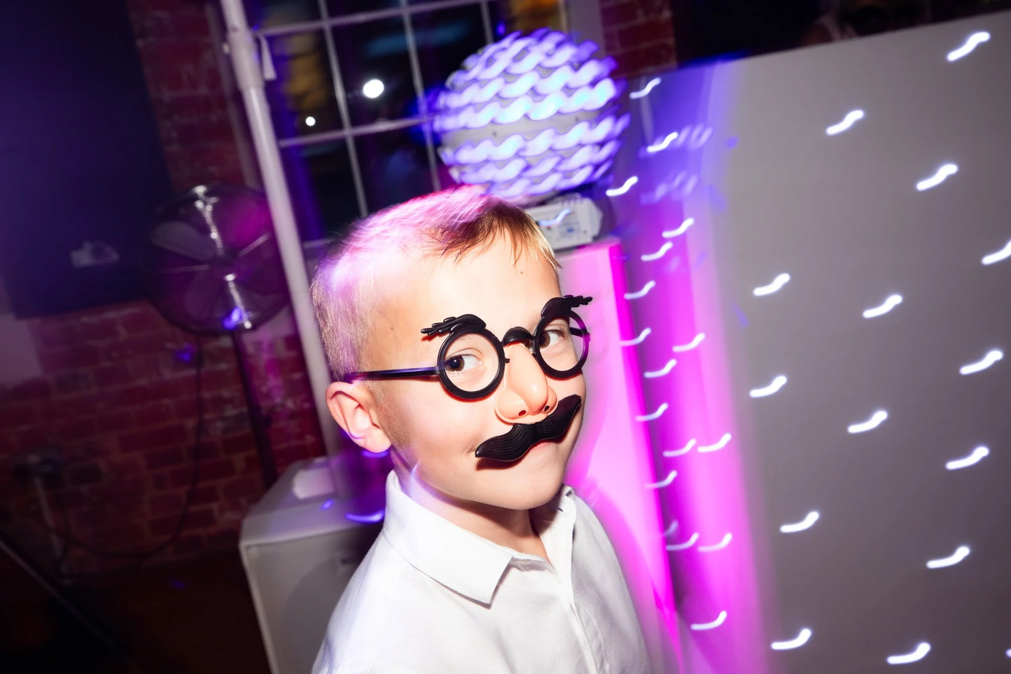 A young boy wearing novelty glasses with a fake mustache and a big nose, looking at the camera with a playful expression. The background features colorful lights and a decorative spherical lamp.