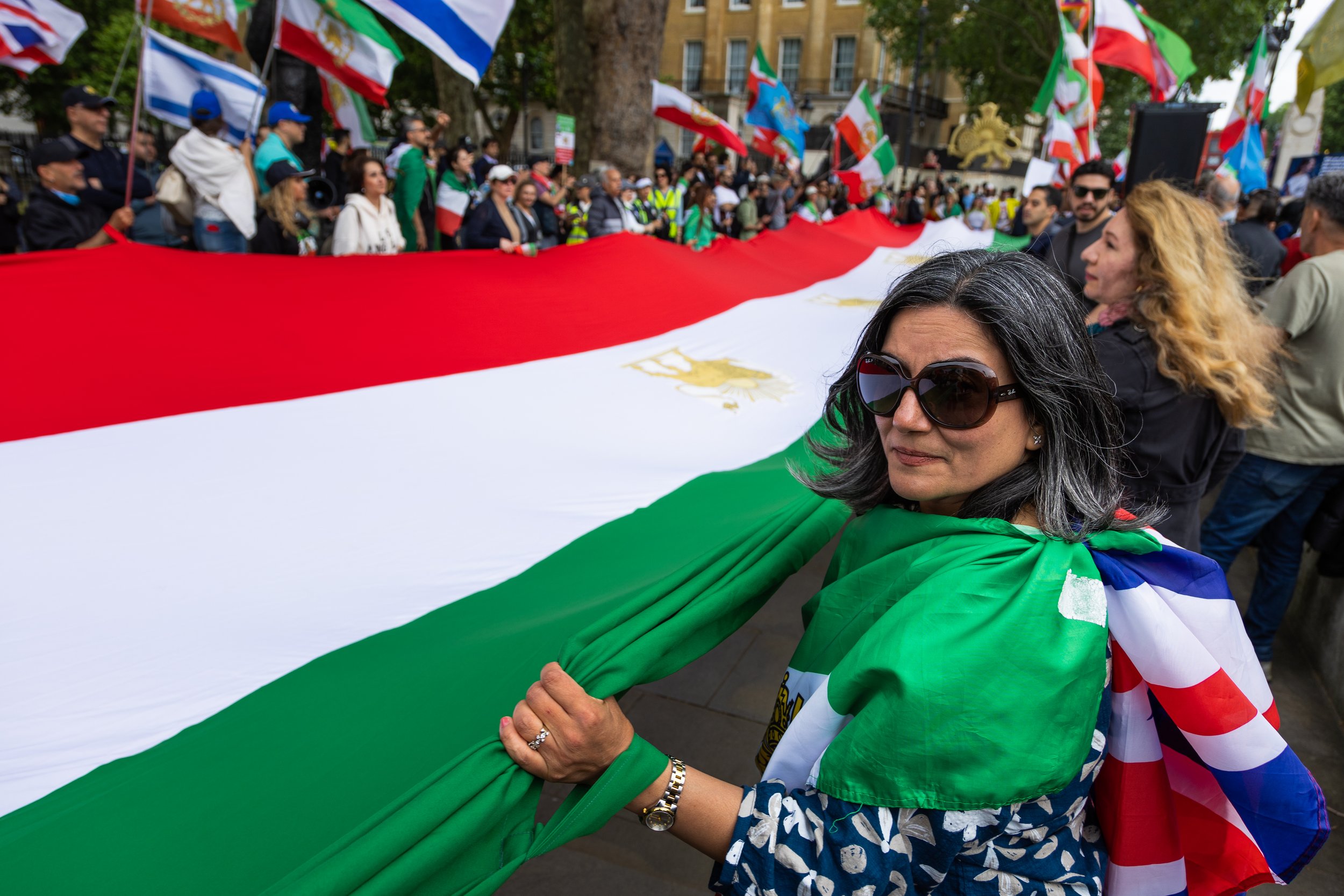 Group of people holding and displaying a large flag with the Mexican emblem, participating in a public demonstration or celebration outdoors with flags from multiple countries in the background.