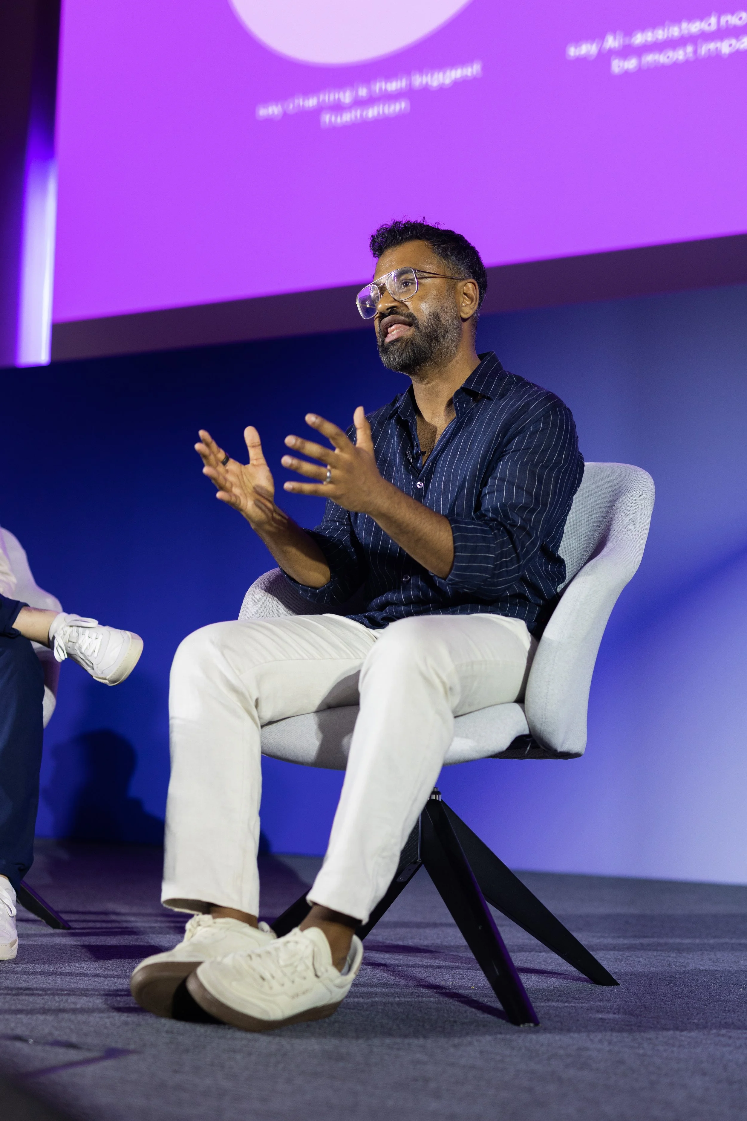 A man with glasses, a beard, and dark curly hair, wearing a black striped shirt and white pants, sitting on a gray chair on a stage, speaking or explaining with hand gestures. There is a purple presentation screen behind him.