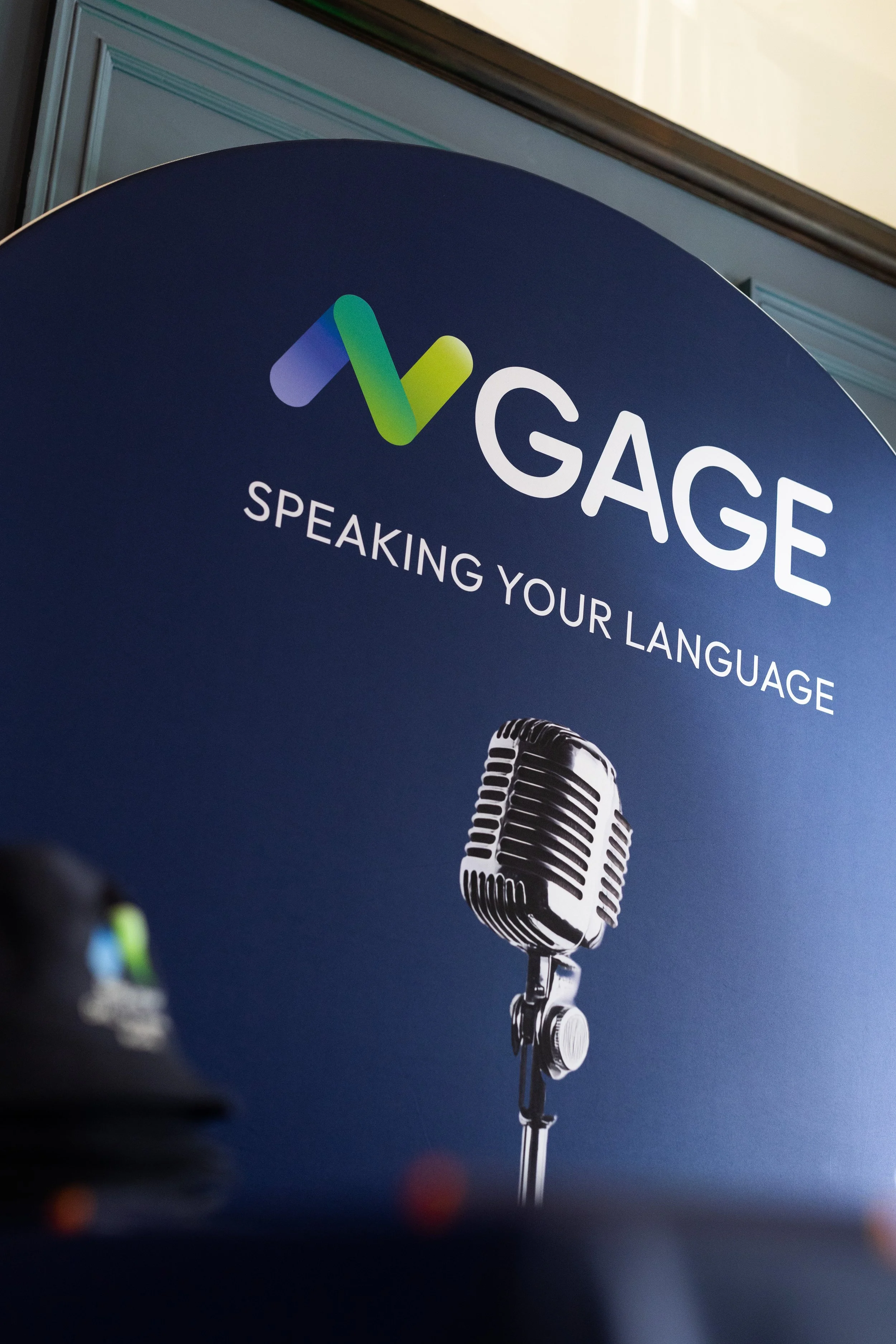 A promotional sign with a logo and microphone graphic, displaying the text 'GAGE SPEAKING YOUR LANGUAGE'.